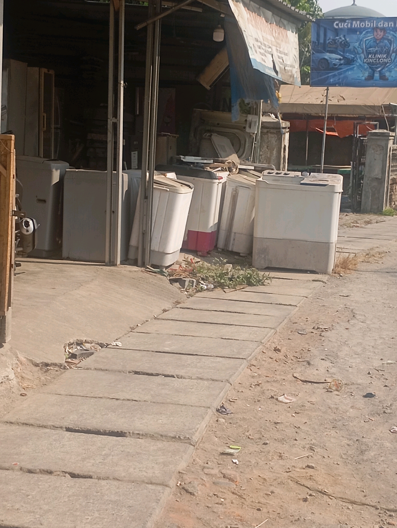 The image depicts an outdoor scene in front of a business establishment in Babelan, Indonesia. The foreground features a cracked concrete pathway with several rectangular concrete slabs. Beyond the pathway, there is a dirt area scattered with debris. In the middle ground, a row of washing machines, some with visible red and white accents, are lined up in front of a covered area. The structure appears to be a simple open-fronted shop with a corrugated metal roof, partially obscured by a tattered banner. A sign on a blue background, with text including "CUCI MOBIL dan," is visible on the right side of the frame, suggesting a car wash service. The background shows a building with a domed roof and other structures. The overall impression is that of a commercial area with an emphasis on appliance sales or repair, set against a backdrop of urban infrastructure.