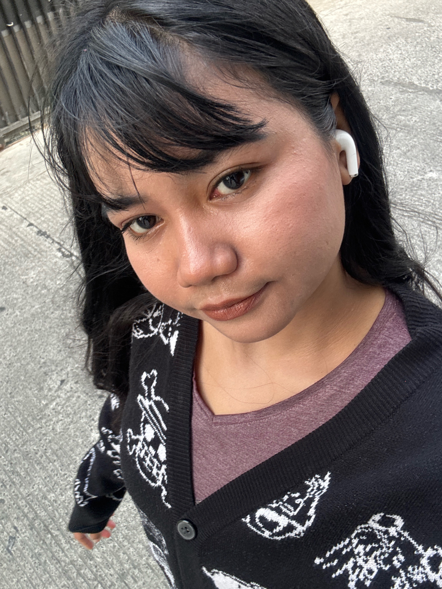 A young woman with black hair and a black and white patterned cardigan stands outside on a concrete surface, possibly a sidewalk or road. She is wearing white wireless earbuds in her right ear and a purple shirt underneath her cardigan. Her gaze is directed slightly upward and to the left, and her lips are closed, with a hint of a smile. The background is slightly blurred, showing a hint of a fence or railing on the left side. The lighting suggests it is daytime. The location is North Jakarta, Indonesia.