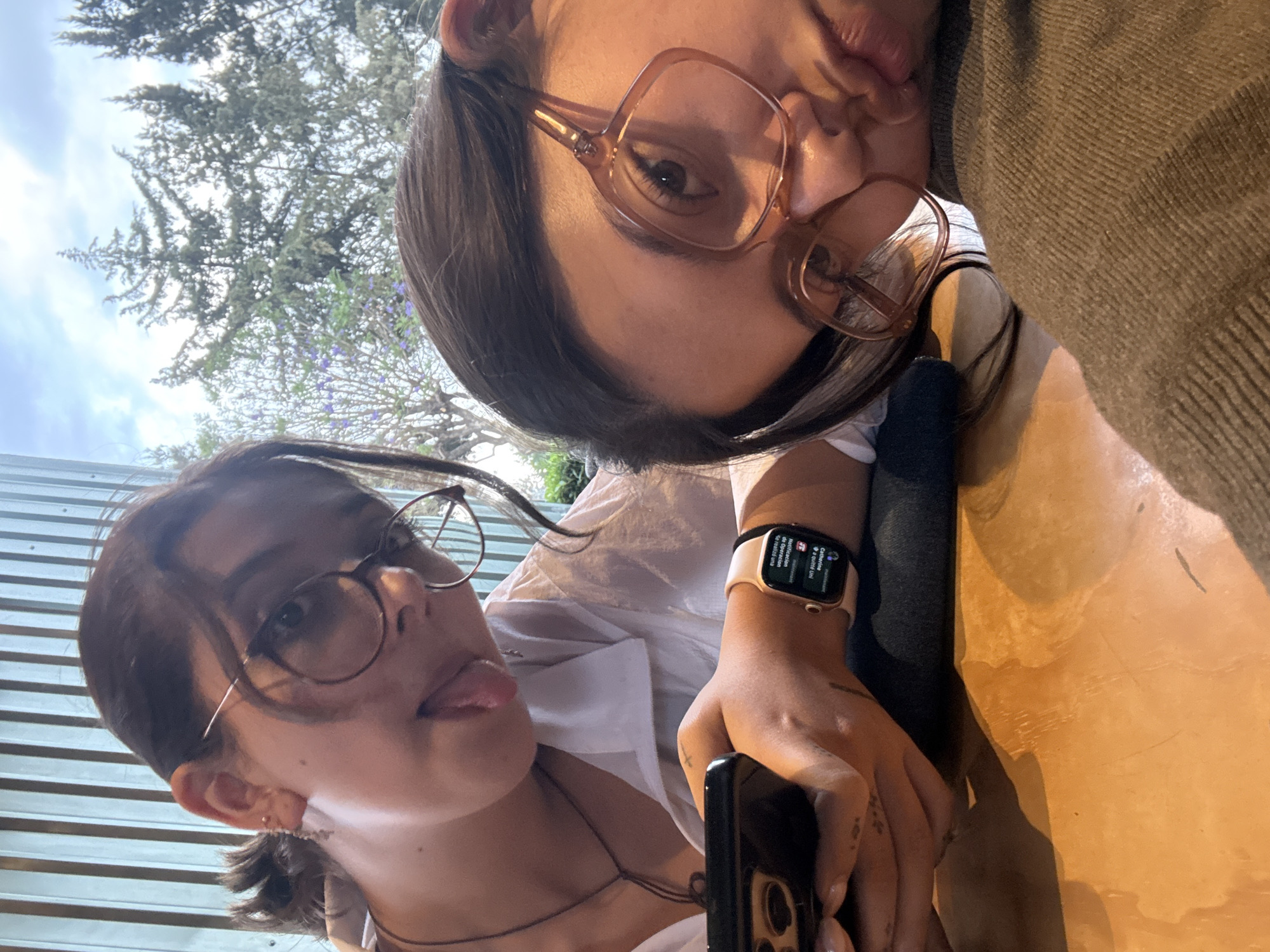 Two young women pose for a selfie. The woman on the left wears eyeglasses and has her tongue sticking out slightly. She is dressed in a white shirt and has a necklace. Her left hand holds a dark-colored smartphone, and she wears a gold-colored Apple Watch on her left wrist. The woman on the right wears oversized eyeglasses and puckers her lips as if giving a kiss. She is wearing a grey sweater. They appear to be sitting at an outdoor table, as suggested by the wooden surface and corrugated metal wall in the background. The sky and trees are visible above them. The photo was taken outdoors in the city of San Andrés Cholula, Mexico.