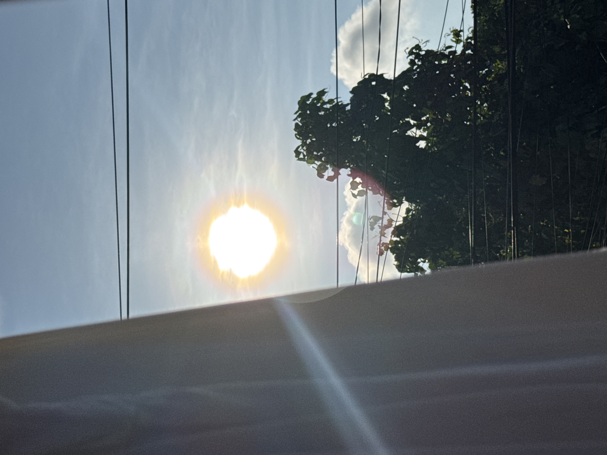The image, taken from a low angle, shows a bright sun in a slightly cloudy blue sky. Several electrical wires are visible, running vertically across the frame. To the right, dense foliage of a tree is present, partially obscuring the sky. In the foreground, there is a light-colored, possibly white or grey, flat surface, possibly a wall or roof, with a glare. Without further context, location in Davao City, Philippines is unconfirmed.