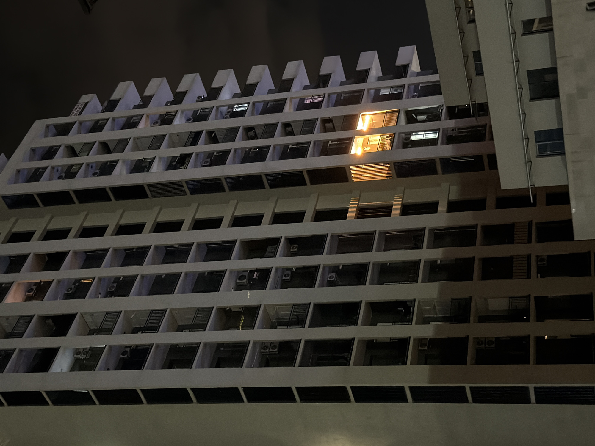 The image shows a tall, modern apartment building at night in Tbilisi, Georgia. The building has a white facade with multiple balconies on each floor. Many windows are dark, but some show interior lights, creating a warm glow. On the right side of the building, a few balconies are illuminated with a bright, yellowish light, suggesting activity within those units. The sky is dark, indicating it is nighttime, and the overall scene is one of urban living. The building appears to be constructed with a contemporary architectural style, featuring clean lines and a repeating pattern of balconies and windows.