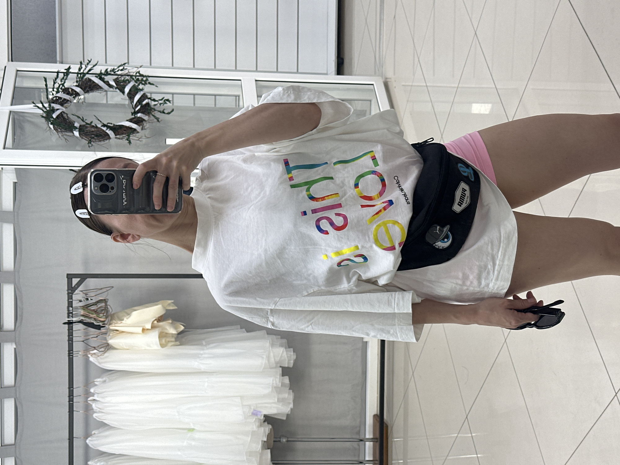 A woman takes a mirror selfie using a smartphone in a room with white tiled floors. She has brown hair with two hair clips, wearing a white t-shirt with colorful lettering, a black fanny pack, and pink shorts. She is holding sunglasses in her left hand. Behind her is a clothing rack with white garments covered in plastic. A wreath hangs on the window. This scene is not necessarily in Votkinsk, Russia, and the location cannot be verified from the image.