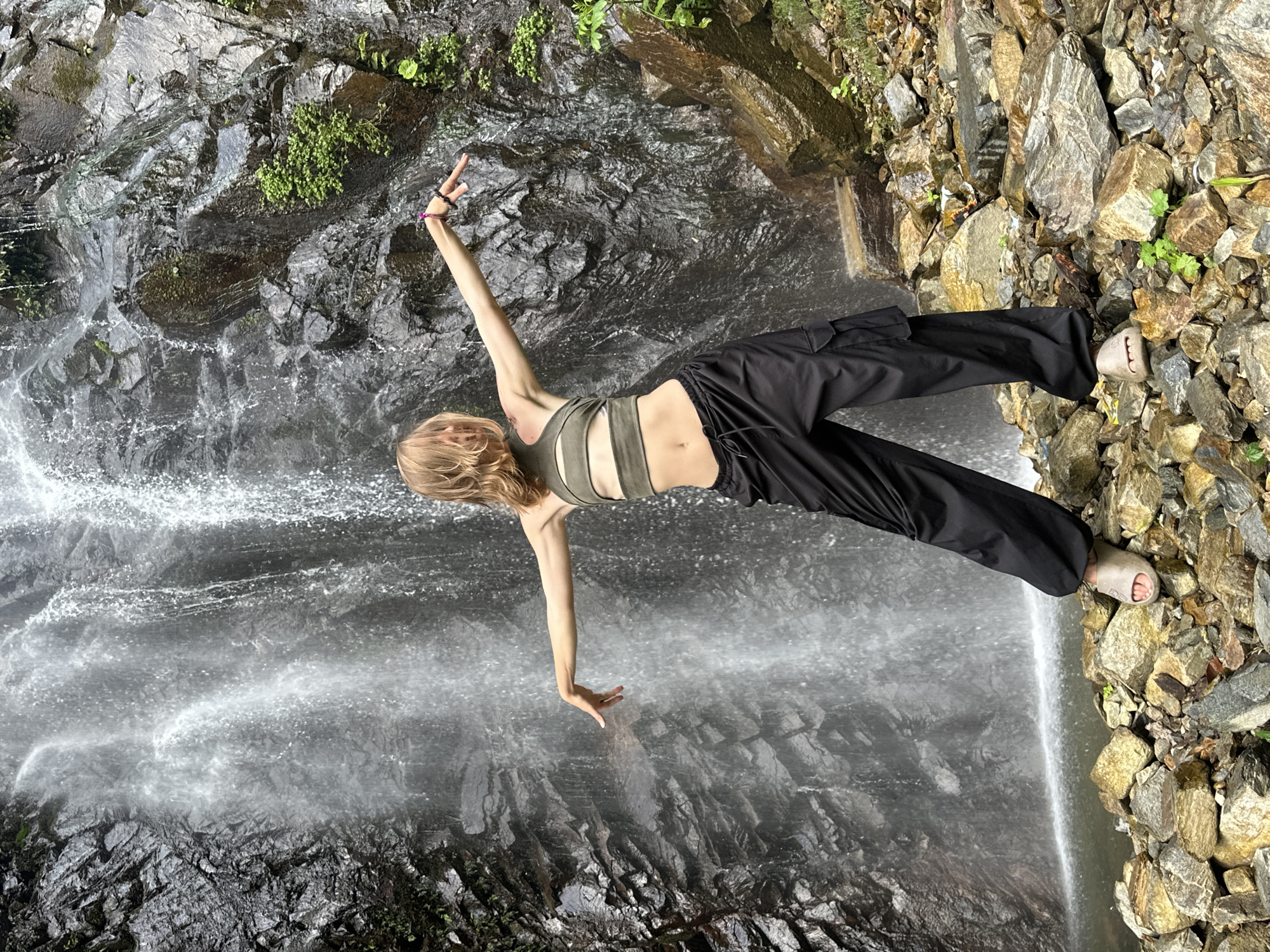 The image depicts a young person posing in front of a waterfall in Tu Le, Vietnam. The subject, a female with blonde, wavy, shoulder-length hair, is positioned centrally, facing away from the camera and slightly to the left. She wears an olive-green crop top with horizontal cut-outs across the midriff and loose-fitting black cargo pants. Light beige open-toed slide sandals cover her feet. Both arms are extended horizontally outwards, with hands slightly cupped downwards; her right arm is slightly higher and more extended than her left, adorned with multiple dark and one pink bracelet near the wrist. The waterfall behind her features a significant volume of water cascading down a dark, jagged rock face, forming a white, frothy pool at the base. Patches of green vegetation are visible on the wet rocks surrounding the waterfall. The foreground consists of an uneven terrain composed of various sizes of wet, multi-colored rocks (grey, brown, yellow), with sparse green plant growth scattered among them. The scene is illuminated by natural daylight, suggesting an outdoor environment.