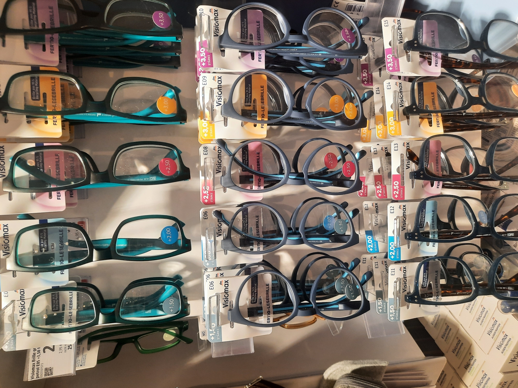 The image shows a retail display of "Visiomax" reading glasses in a store, likely a drugstore or a department store, in Rosenheim, Germany. The glasses are arranged in rows and columns, with each pair packaged in plastic and cardboard. The packaging indicates the strength of the lenses, with various diopter values such as +1.50, +2.00, +2.50, +3.00, and +3.50. Some packaging also highlights that the glasses are made from 95% recycled material. The overall environment appears to be brightly lit and organized, typical of a retail setting. There are no people visible in the image, and the focus is entirely on the product display. The background is a plain white wall, suggesting a clean and modern store design. The products are predominantly framed in black and blue, with some green frames also visible. The price labels are predominantly white with text in black and blue.