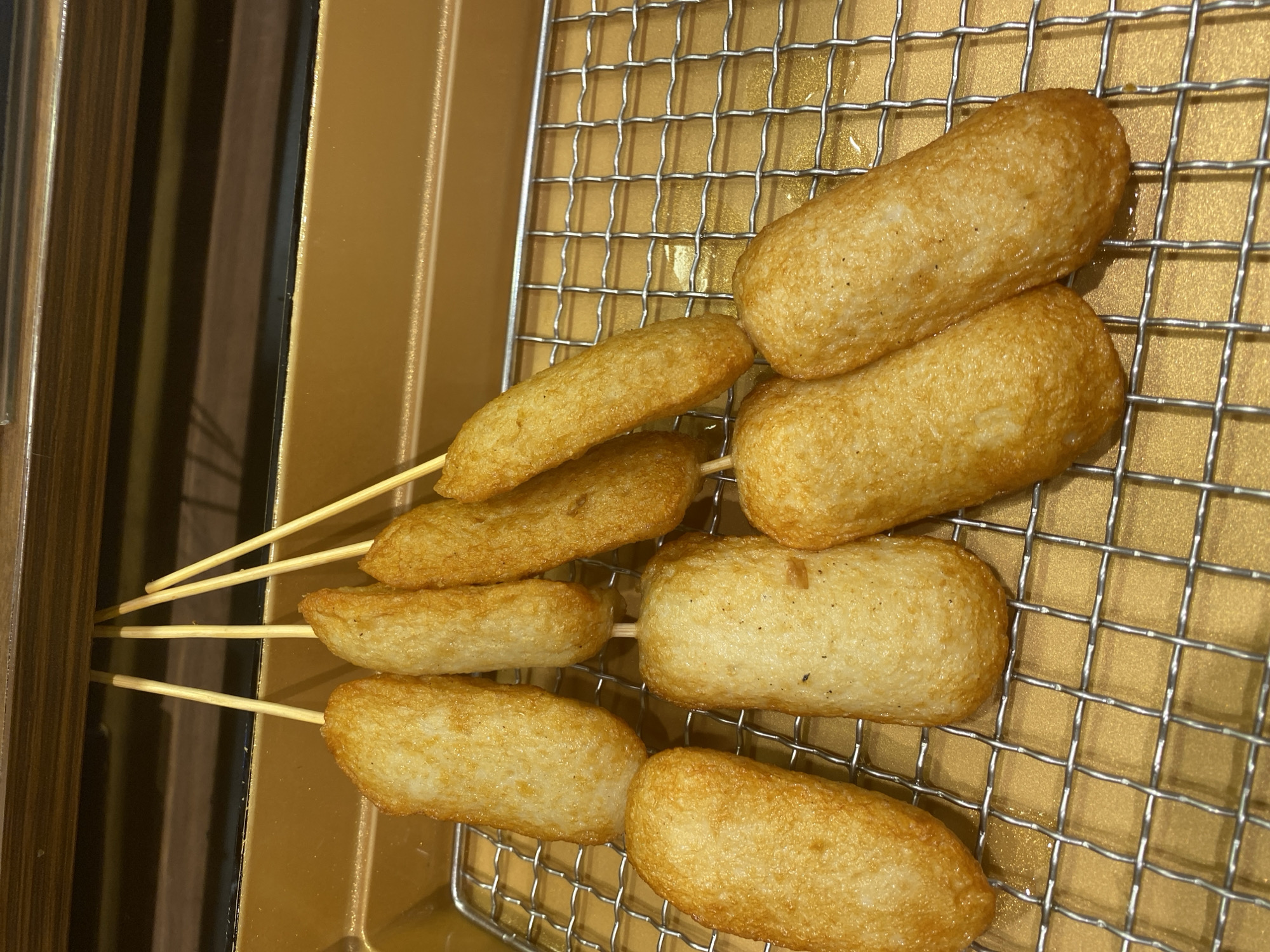 A close-up, top-down shot shows eight fish cakes on wooden skewers, arranged on a wire rack. The fish cakes are golden brown and appear to be fried. They are oval or cylindrical in shape and are of varying sizes. Some skewers hold one fish cake, while others hold two. The wire rack sits on a golden-colored surface, which appears to be a serving tray or a part of a kitchen appliance. The background is a blurred wooden surface. The scene is likely indoors, possibly in a restaurant or food stall. This image was taken in East Jakarta, Indonesia.