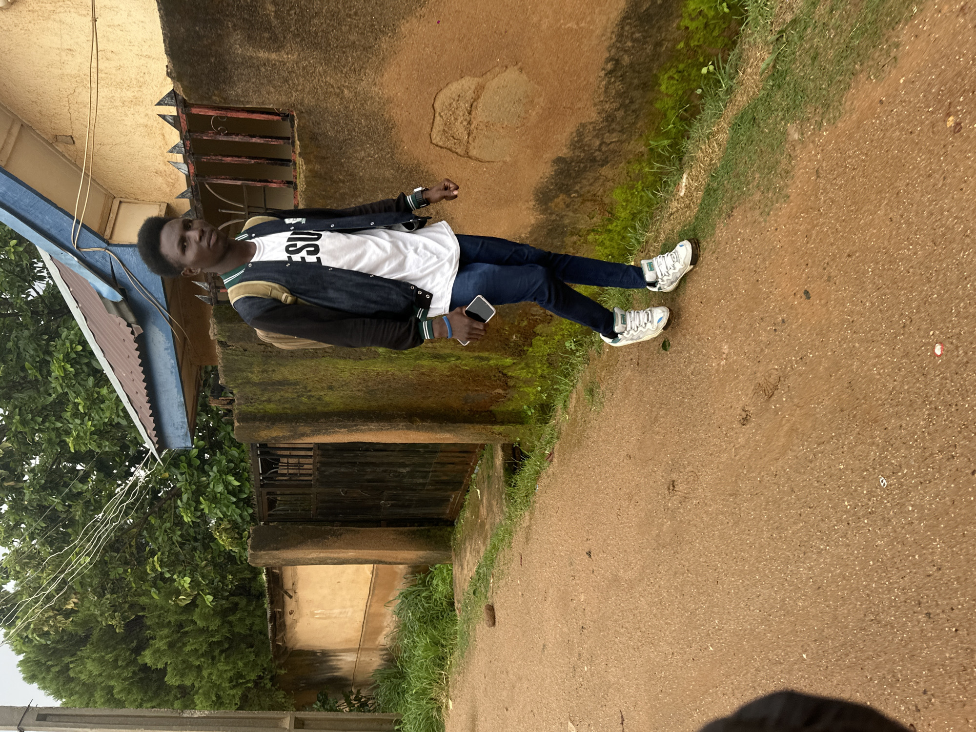 The image shows a young man standing outdoors in what appears to be a residential area. He is wearing a white t-shirt with black text that reads "JESU", dark blue jeans, and white sneakers. He is also wearing a dark jacket with green and white accents. He has a backpack slung over one shoulder and is holding a smartphone in his right hand.
The setting is characterized by a building with a yellow-brown textured wall and a blue corrugated metal roof extension. There is a barred window on the building, and moss is visible on parts of the wall. To the left of the building, there is lush green vegetation, including trees and grass. The ground is a reddish-brown, gritty surface, suggesting it could be a dirt road or pathway. A patch of grass lines the edge of the pathway.
The image is taken from a slightly elevated perspective, looking down at the man. The lighting suggests it is daytime, and the overall atmosphere appears casual and everyday. The man's posture and gaze do not convey strong emotions, but he appears relaxed.
Visible text: "JESU" on the t-shirt.
Location context: Gwong, Nigeria.