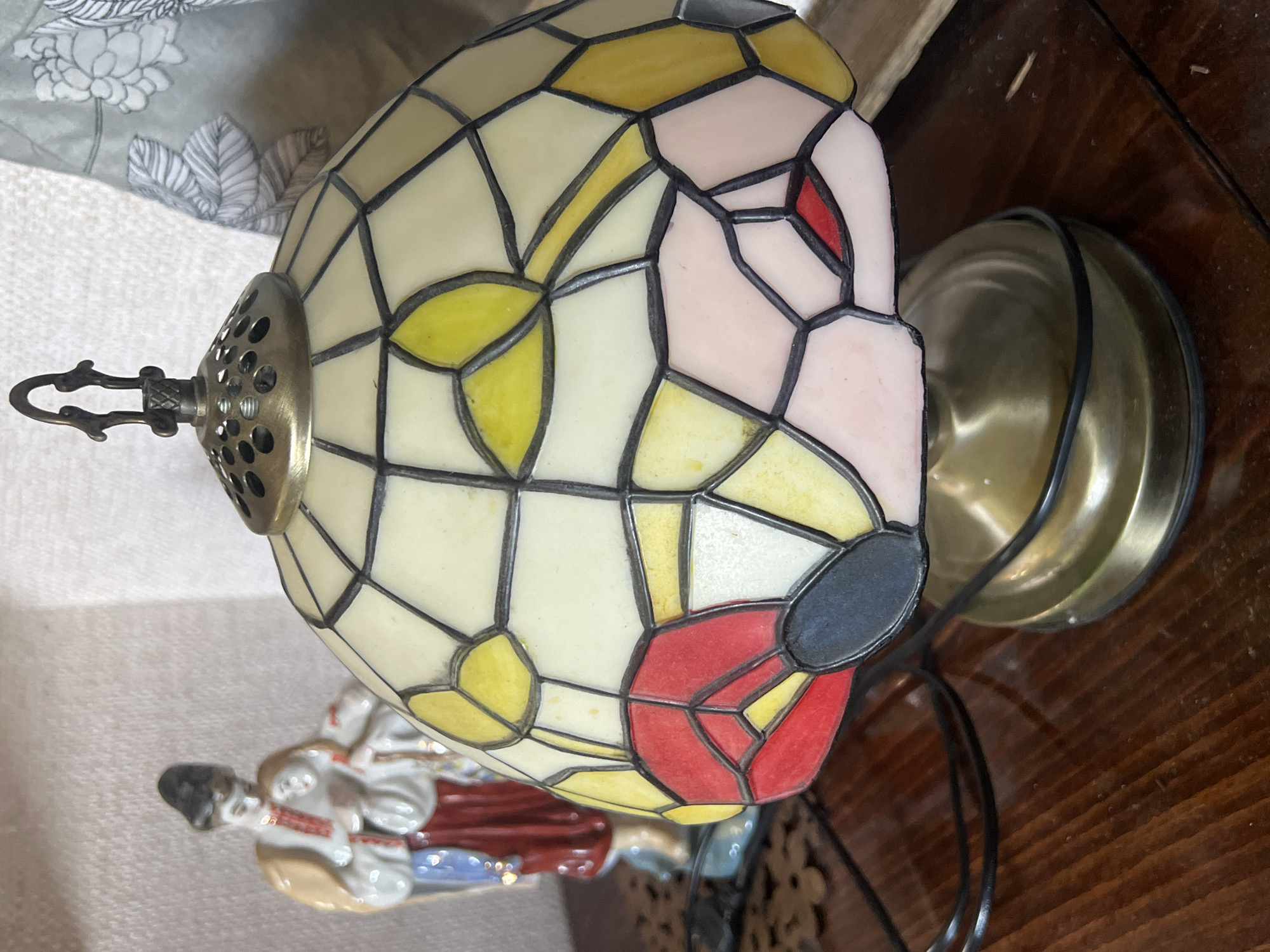 The image shows a close-up of a stained-glass table lamp with a Tiffany-style lampshade. The lampshade is made of various colored glass pieces, including yellow, pink, red, and white, with black lead lines connecting them. The lamp has a brass base and a decorative metal finial at the top. Behind the lamp, a small porcelain figurine of a woman in traditional clothing can be seen. The figurine is wearing a red skirt, a white blouse with red embroidery, and a blue sash. She is holding a baby. The objects are placed on a wooden surface, likely a table or shelf. The background is blurred, but appears to be a wall and some fabric with a floral pattern. The scene is indoors, and the lighting suggests it might be daytime, with artificial light from the lamp also possibly contributing. The specific location in the city of Norviliškės, Lithuania is not ascertainable from the image itself.