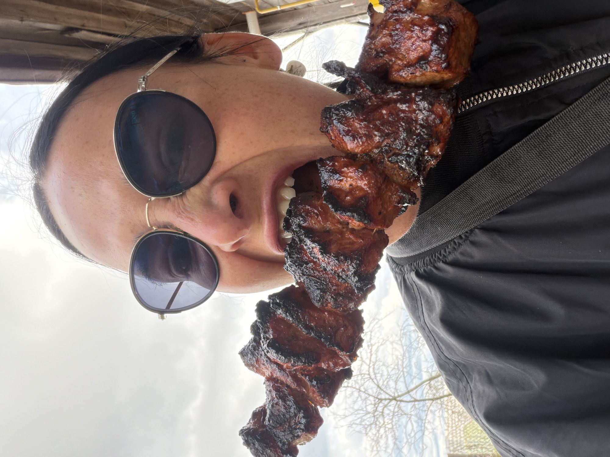 A person wearing round sunglasses and a black jacket holds a skewer of cooked meat up to their mouth. The person has a nose ring in their left nostril. The environment appears to be outdoors, potentially in Бронница, Russia, given the prompt's instruction, with a view of the sky and some trees visible in the background.