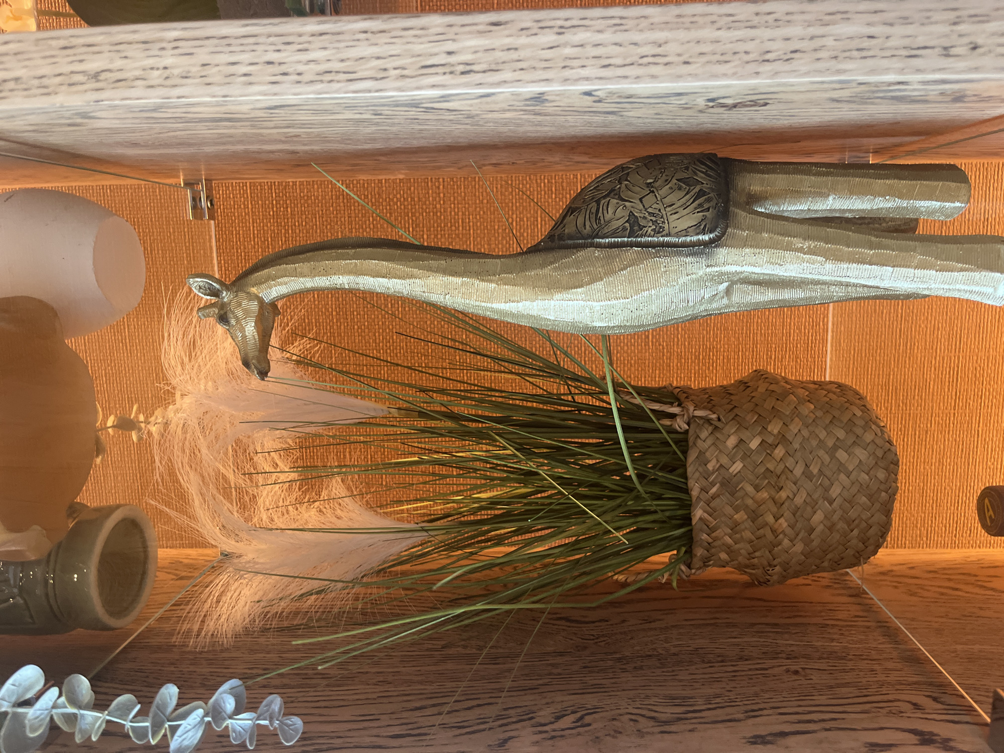 The image displays a still life arrangement of decorative objects inside a shelving unit. The primary subject is a tall, slender giraffe sculpture, light brown with subtle stripes and a patterned saddle-like detail on its back. The giraffe's head faces towards the left, and it appears to be standing upright on a wooden shelf.
Next to the giraffe, a woven straw basket contains a bundle of long, green artificial grass and several fluffy white plumes, resembling pampas grass. Further to the left, on the same wooden shelf, are a smooth, off-white, egg-shaped decorative item and a light green ceramic vase or container, which is lying on its side. In the foreground, a branch with pale, oval-shaped leaves, similar to eucalyptus, is partially visible.
The objects are placed against a textured, orange-brown back wall, which appears to be wallpaper or fabric. The shelves themselves are made of light-colored wood with a noticeable grain. A small, silver metal bracket is visible connecting one of the shelves. No activity is taking place, as it is a static display. There is no clearly discernible text within the image. The scene suggests an indoor decorative display, possibly within a home or commercial setting.