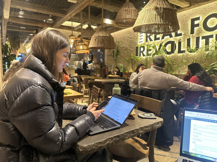 The scene is set inside a modern cafe or restaurant, strongly indicative of a "Healthy Poke" establishment, a "Real Fooding" concept eatery located in Madrid, Spain. The interior features a rustic yet contemporary design with warm, indirect lighting provided by multiple large, woven pendant lamps suspended from a ceiling constructed with wooden beams. A prominent textured wall, in a light, neutral tone, displays illuminated text that reads "...THE REAL FOOD REVOLUTION," partially obscured, and is decorated with several green potted plants.
In the foreground, to the left, a female with long, dark brown hair, wearing a black puffer jacket, is seated at a dark, irregularly shaped stone or concrete table. She is actively engaged with a black smartphone, holding it in her right hand and looking at its screen. A silver laptop, displaying lines of text, is open on the table in front of her, with her left hand resting near its keyboard. Also on her table are a light green reusable water bottle and a grey circular coaster or charging pad. To the immediate right, another silver laptop, open and displaying text, is partially visible.
In the midground, to the right, a man with short dark hair, wearing a light brown turtleneck sweater, is seated with his back mostly to the viewer, his right arm extended towards a woman seated to his right. This woman has long dark hair, wears a red top, and appears to be in conversation with the man, with her left hand resting near her chin. Another woman, wearing a light-colored top, is partially visible to the man's left. Several other patrons are visible in the blurred background, seated at various wooden tables, and one individual is seen near what appears to be a service counter or open kitchen area. The overall ambiance is casual, suggesting a place for dining, socializing, and working.