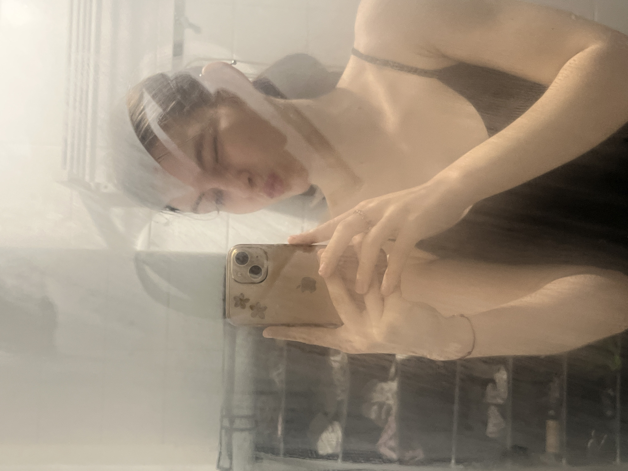 A young woman is taking a selfie with a gold-colored iPhone. Her face is slightly blurred due to the lighting and the phone's proximity. She wears a thin-strapped garment, possibly a tank top or dress. Her hair appears to be pulled back with a hairband or clip. A ring adorns one of her fingers. The setting seems to be a bathroom, indicated by the hazy, mirrored or reflective surface in front of her. There is no evidence to support that this image was taken in Saint Petersburg, Russia.