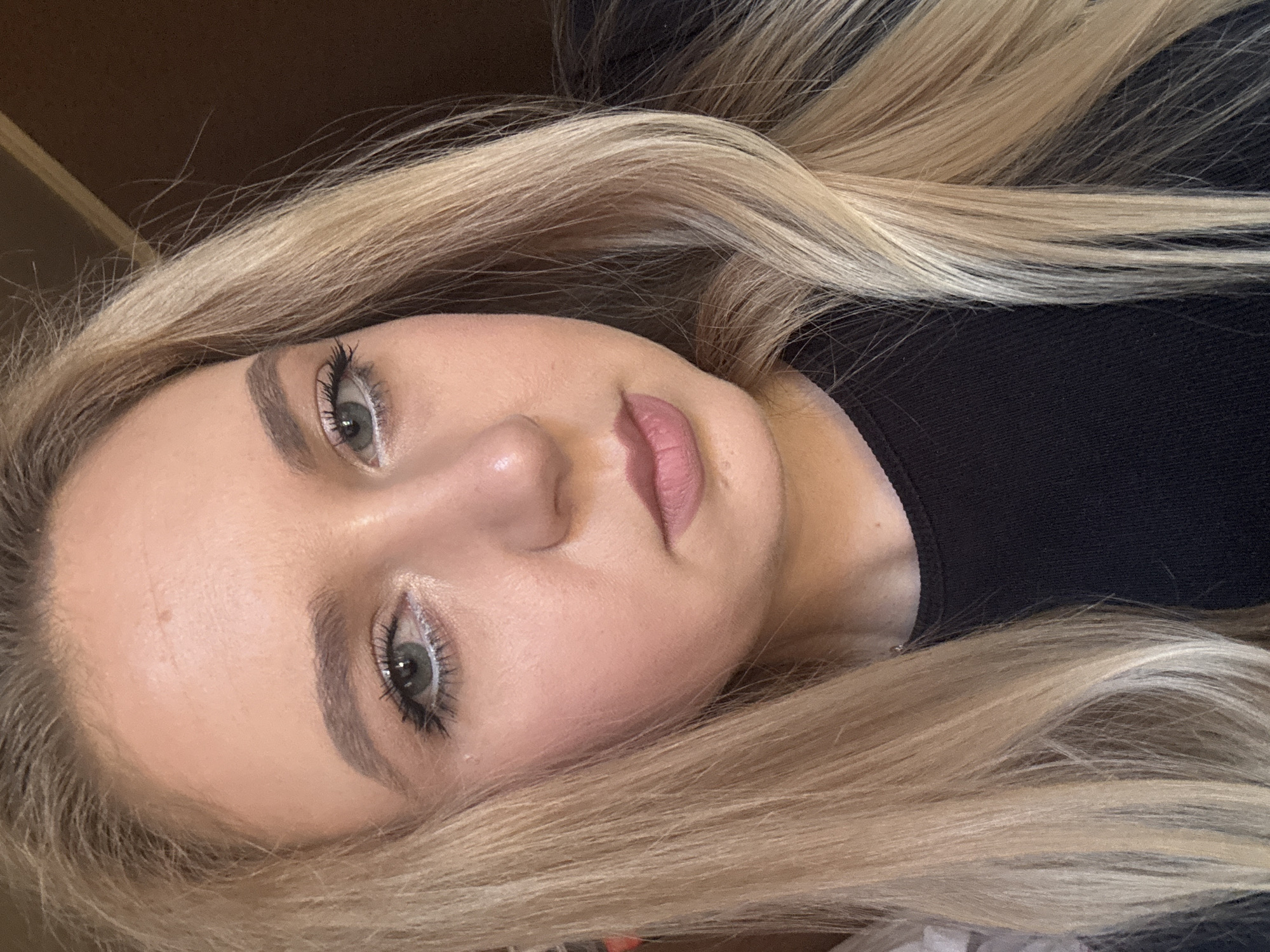 This is a close-up, chest-up, portrait-style photograph of a young Caucasian woman with long, straight blonde hair. She is wearing a black top and her head is slightly tilted to the right. Her left eye is looking directly at the camera, while her right eye is partially visible. She has light-colored eyes with visible mascara on her lashes and white eyeliner on her lower lash line. Her eyebrows are well-defined and filled in. Her skin appears fair with a slight blush on her cheeks. She is wearing a matte, pinkish-brown lipstick. The background is a dark brown color, suggesting an indoor setting. The lighting is warm and appears to be coming from the left side of the frame, creating some highlights on her face and hair. There is no indication of the location being in a city, as it appears to be a neutral, indoor environment.