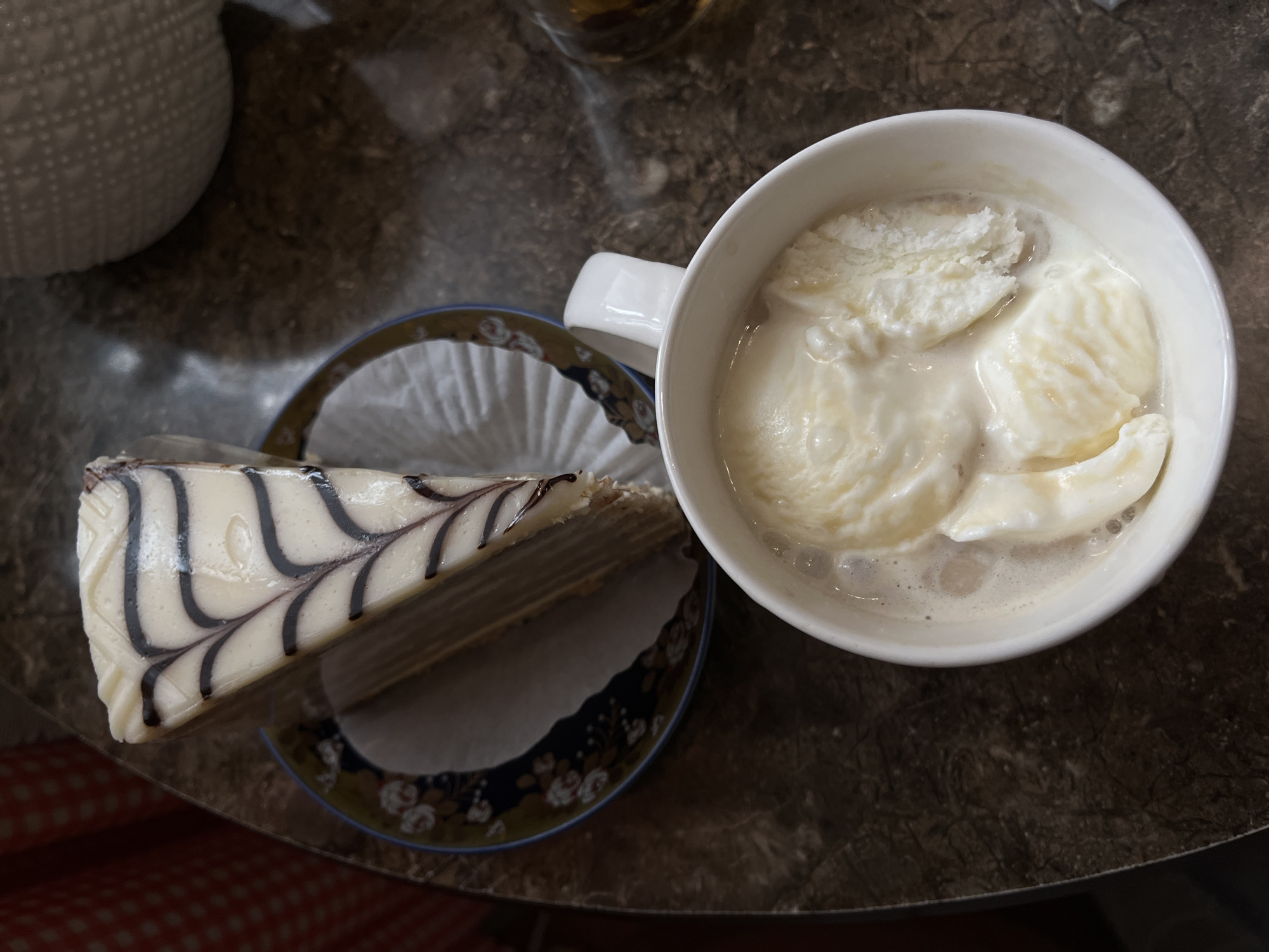 A slice of vanilla cake with dark chocolate drizzle, presented on a decorative plate, sits next to a white mug filled with melted vanilla ice cream. The scene is captured from a top-down perspective, with the dark, speckled surface of a table as the backdrop. The lighting suggests an indoor setting, possibly a cafe, and no individuals are visible. The location of Petropavlovsk-Kamchatsky, Russia, is not ascertainable from the visual information provided in the image.