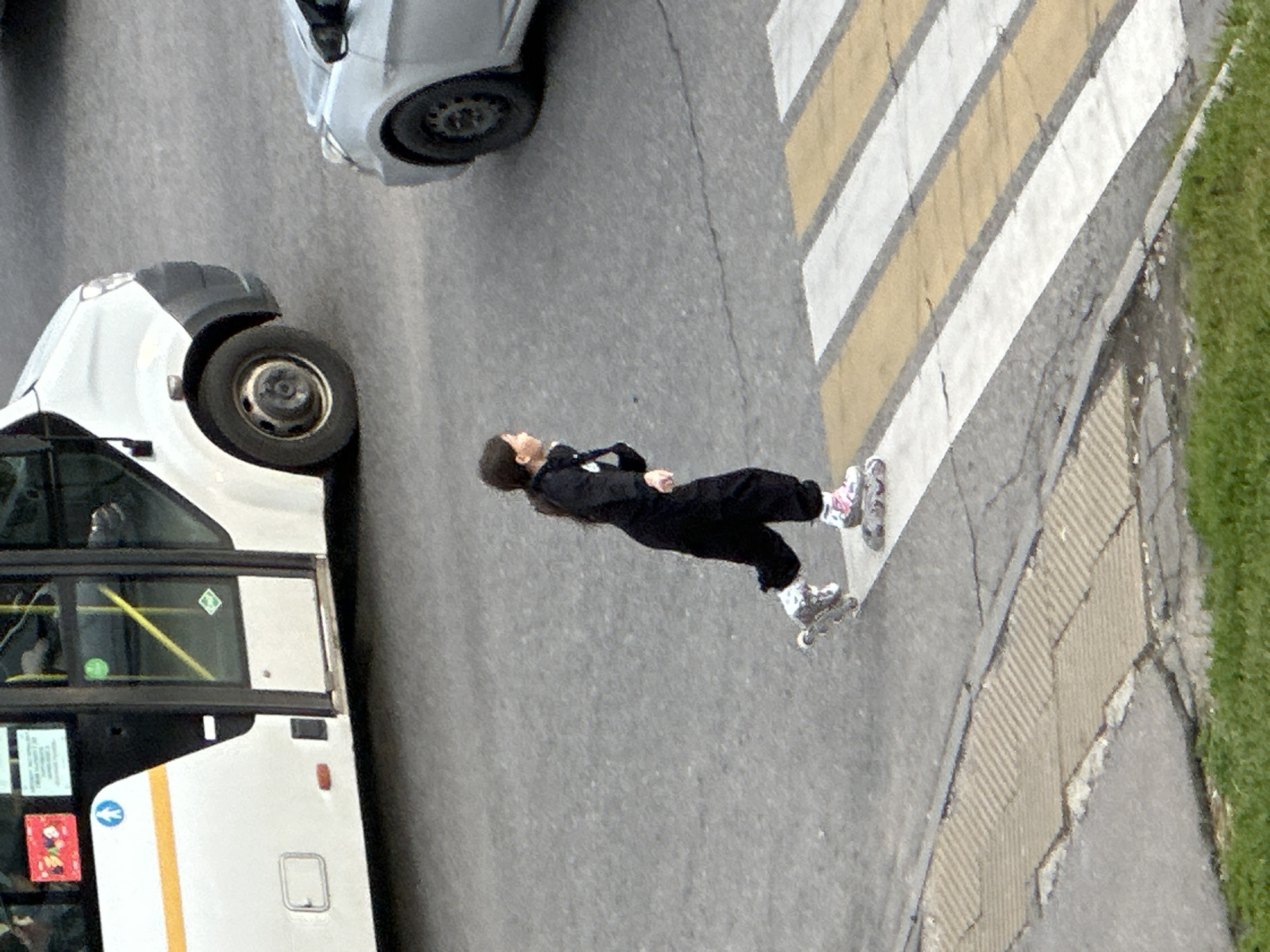 In Podolsk, Russia, a young person in black athletic wear and roller skates is captured mid-stride, crossing a crosswalk. The individual's body is angled slightly to the left, with their right leg extended forward and their left leg trailing. They are wearing white and pink roller skates. Behind them, the distinct yellow and white stripes of the pedestrian crossing are visible, along with a silver car stopped to their right. Further in the background, the side of a white bus with orange and blue accents is parked, featuring tinted windows and a visible wheel. The ground surface consists of asphalt and a textured grey pavement, with a strip of green grass bordering the bottom of the frame. The overall scene appears to be an urban street environment during daylight hours.