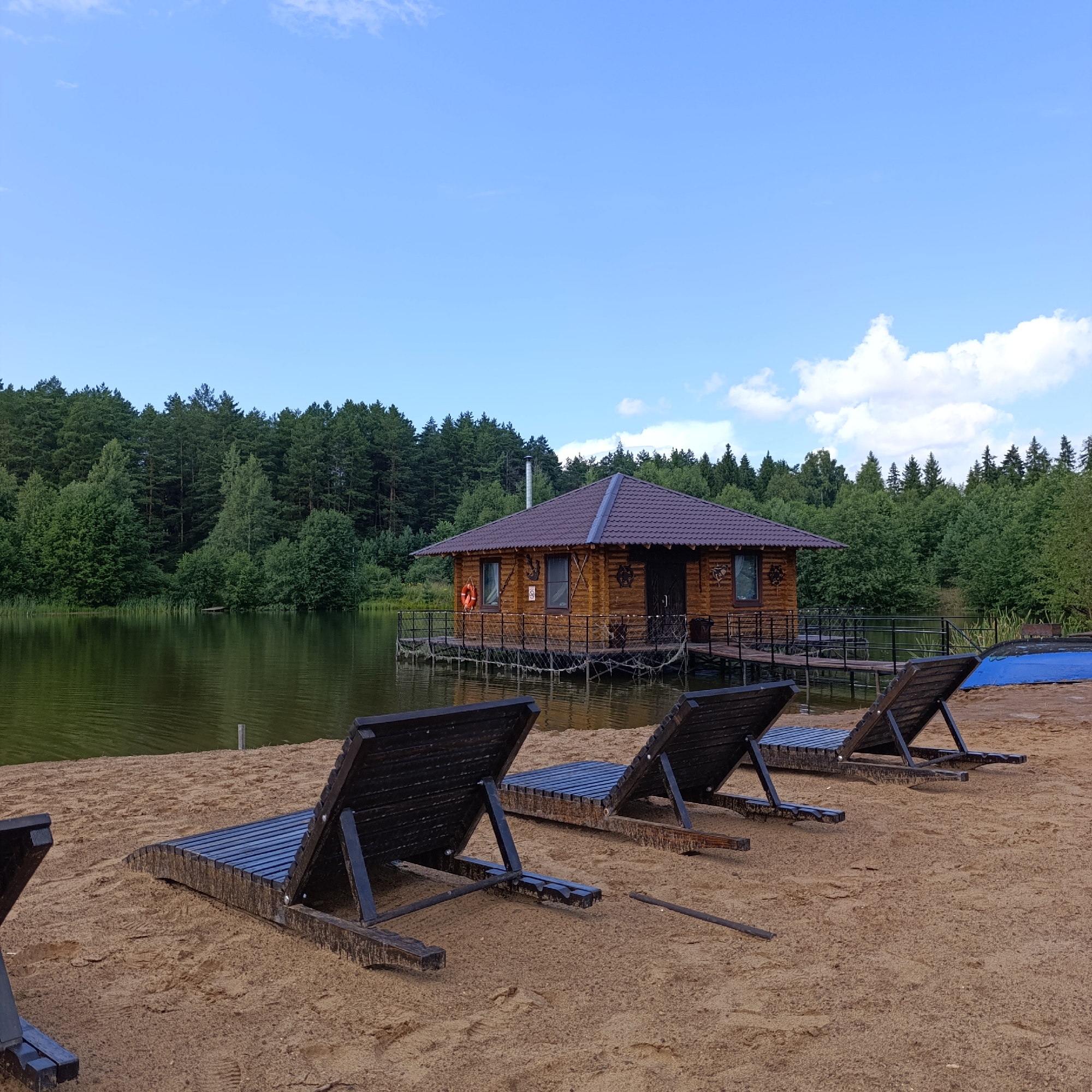 An outdoor scene depicts a tranquil lake bordered by a dense forest of green trees. In the foreground, three dark wooden lounge chairs are set on a sandy beach, angled towards the lake. A wooden structure resembling a small house or sauna is situated on a platform over the water, with a dark-tiled roof and wooden walls. A life preserver ring is visible on the exterior wall of the structure. The sky above is a bright blue with scattered white clouds. The scene appears to be in Prikamskii, Russia, suggesting a recreational or resort setting. There are no people present in the image.