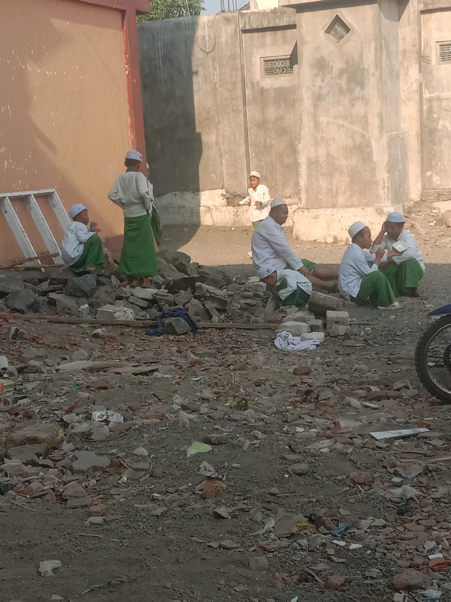 In the city of Sidogiri, Indonesia, several young boys and men, most of whom are dressed in traditional Islamic attire consisting of white shirts and green sarongs, are gathered in an outdoor area. The environment appears to be a construction or demolition site, as evidenced by the abundance of scattered rocks, debris, and rubble. In the background, there is a light brown building with a red frame and a grey concrete structure with a diamond-shaped vent. Some of the individuals are sitting on the rubble, while others are standing or interacting amongst themselves. One boy is crouched down, seemingly examining something on the ground. Another individual, further in the background, is standing near the light brown building. The scene is illuminated by natural sunlight, casting shadows on the ground.