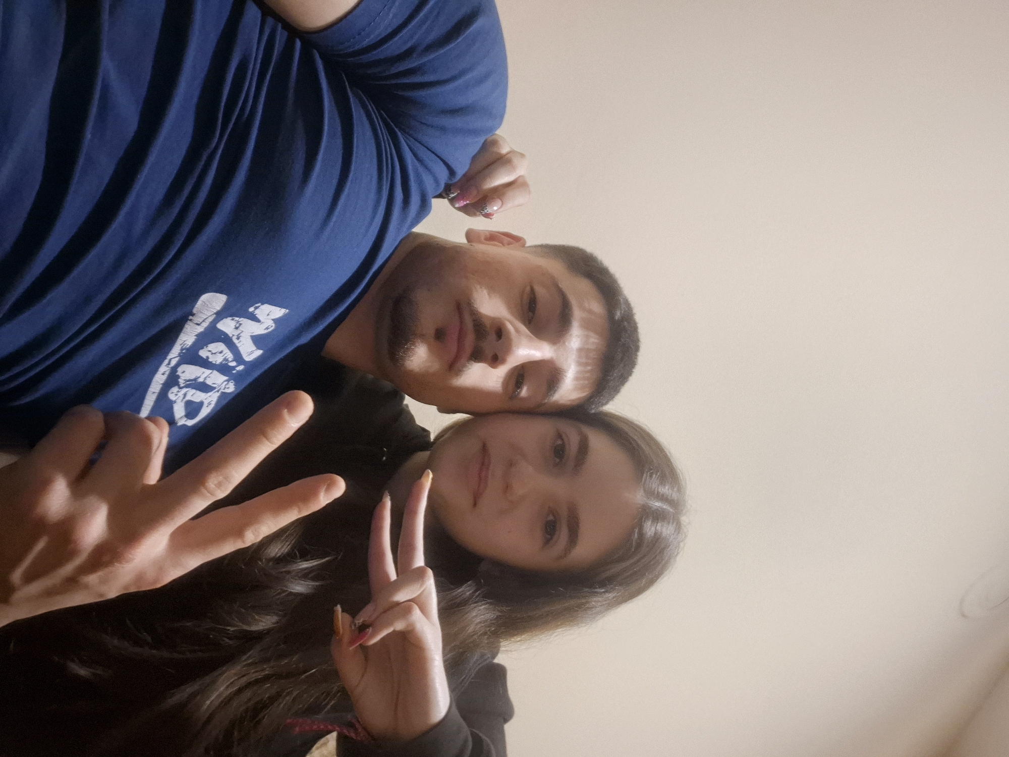 A selfie depicts a man and a woman at an unusual angle from below. The man, positioned at the top of the frame, wears a dark blue t-shirt with a white graphic. The woman, below him, has long brown hair and is wearing a dark hoodie. Both are making the peace sign. The background is a plain, light-colored wall. The location is assumed to be Pavlikeni, Bulgaria.