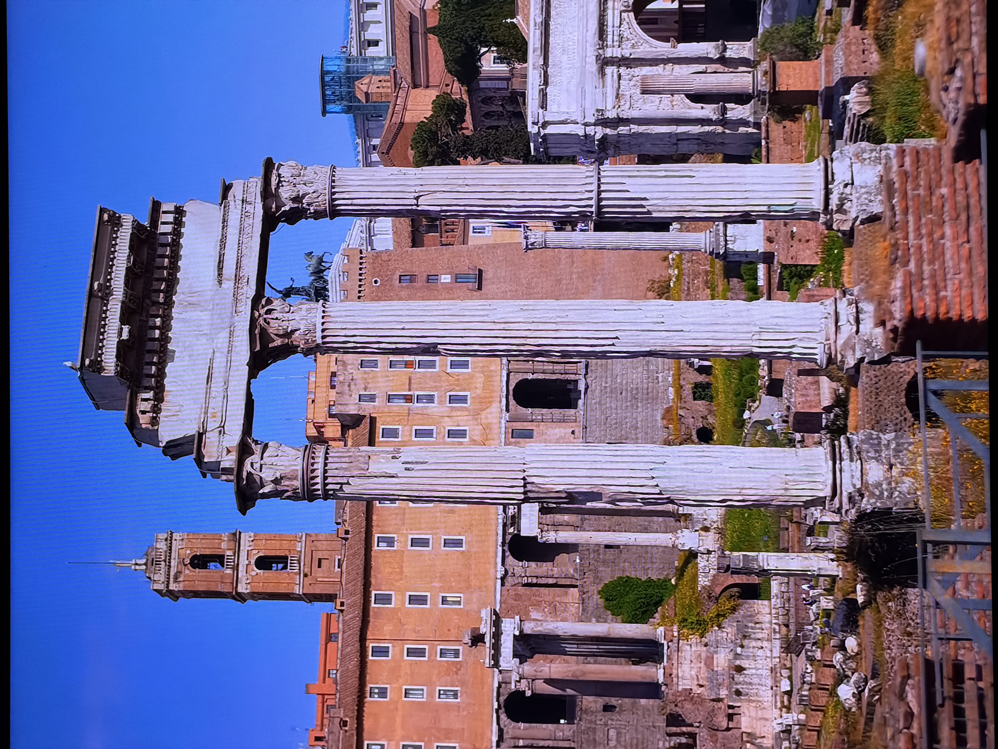 The image shows the ruins of an ancient Roman temple, with tall white columns in the foreground and a clear blue sky in the background. Behind the columns, there are more ruins, including arches and other stone structures. In the distance, there are several buildings with terracotta roofs, and a tall bell tower with an orange facade. There is also a modern glass-enclosed structure visible. The ruins are overgrown with grass and some yellow flowers. A few people are visible in the distance, walking among the ruins. The scene appears to be a historical site under a bright, sunny sky. The city of Chirchik, Uzbekistan, is not depicted in this image.