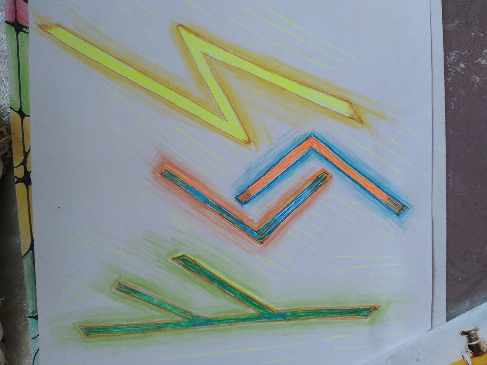 The image shows a close-up of a white sheet of paper with abstract drawings made with colored pencils or crayons. The drawings consist of geometric shapes and lines in green, orange, blue, and yellow. There are no people, objects, or discernible actions visible. The drawing is on a flat surface, and a portion of what appears to be a book or binder with a patterned edge is visible on the right side of the image. The background is a light grey, textured surface, possibly a table. There is no information in the image to suggest a location in the city of Baki, Uzbekistan.
