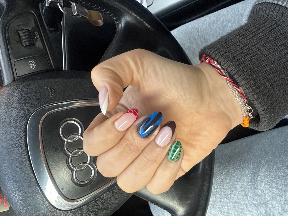 The image presents a close-up of a person's left hand resting on the black steering wheel of an Audi vehicle. The location is Wattrelos, France. The hand displays manicured fingernails with distinct designs: the index finger has a white French tip, the middle finger features a nude base with a red, textured tip, the ring finger is blue with black wavy patterns, and the pinky finger is green with a grid-like texture. The thumb's nail shows a nude base with a dark tip. The person's wrist wears multiple bracelets, specifically a red and white braided string, a clear beaded string, and an orange beaded string with a metallic clasp, emerging from a dark gray ribbed knit sleeve. The steering wheel's center bears a chrome Audi logo, and to its right, vehicle controls and a car key with a brown, possibly wooden, accessory are visible. A red digital display in the upper left of the frame shows "6:02" and "19°". The person's leg is covered by light grey fabric, visible in the lower left.