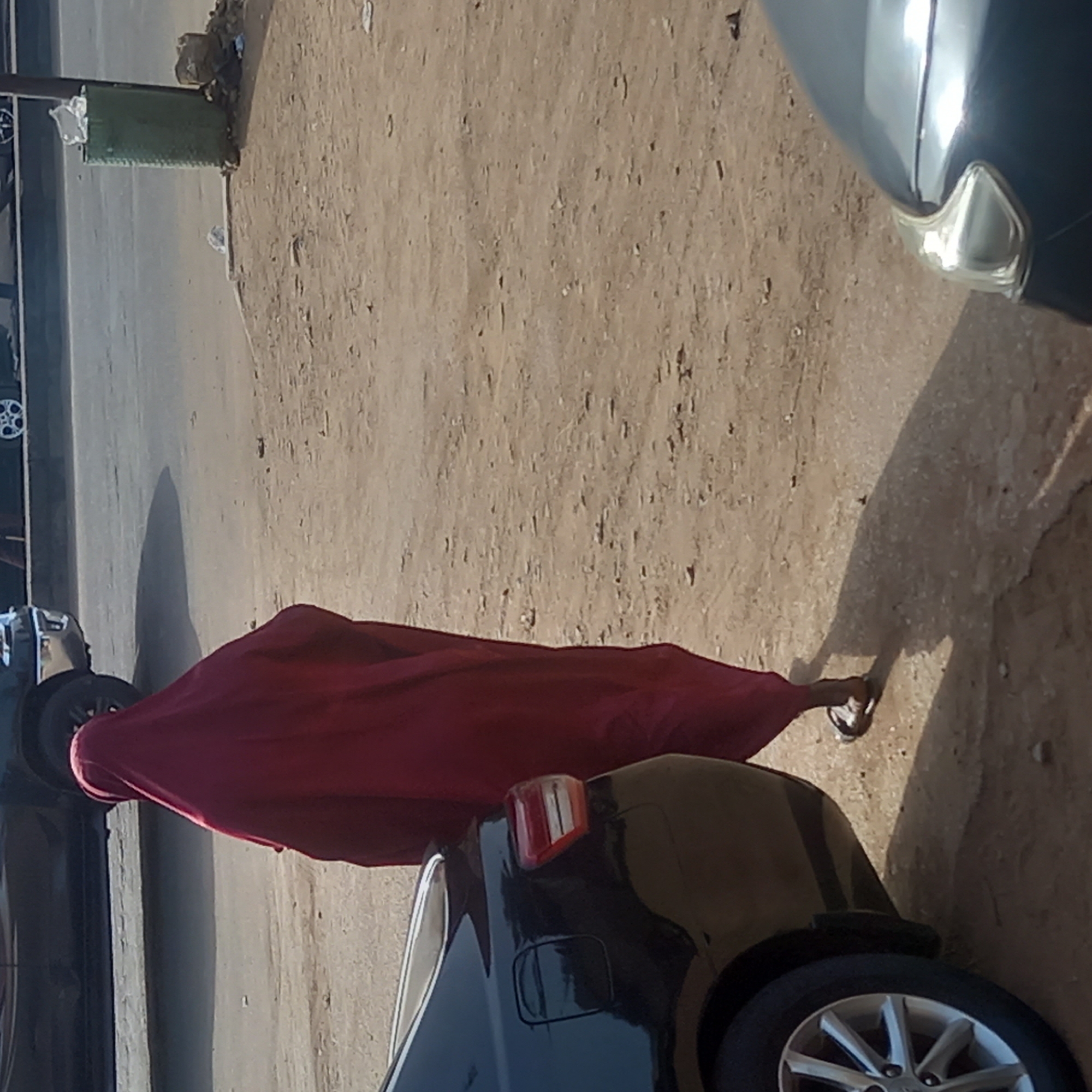 This vertical shot captures a street scene in Dadi Riba, Nigeria, under strong sunlight. The foreground is dominated by a dusty, light brown unpaved area that transitions into a darker, possibly paved road on the left side of the frame.
A central figure, likely a person, walks along this dusty path. They are dressed in a long, flowing crimson red garment that covers their body and head, revealing only their bare feet wearing black and white sandals. A prominent shadow of the person stretches across the ground to their right.
On the far right, partially visible, is the front section of a dark-colored car, showing a portion of its bumper and a clear headlight. In the mid-ground, to the left of the person, is the rear end of another dark-colored car, displaying its tail light, fuel cap cover, and a silver alloy wheel. Another white wheel with spokes is partially visible further in the background on the far left.
Near the left edge of the frame, in the background, stands a rectangular, dark green object, possibly a waste bin, with some trash piled on and around it. The bright, direct light creates harsh shadows and highlights, suggesting a clear, sunny day. No discernible text is visible in the image.