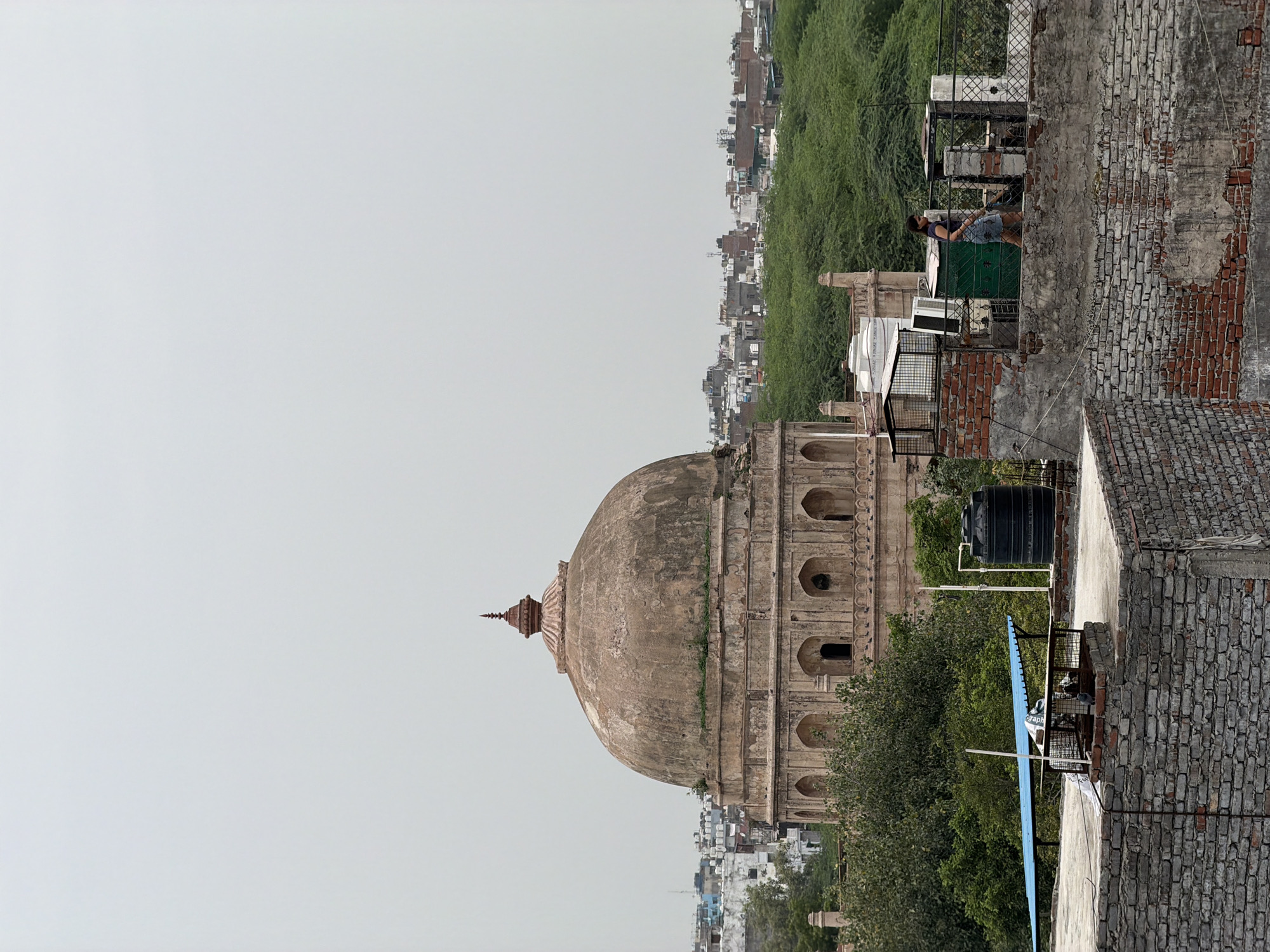 The image shows a view of the Lodhi Garden tombs in Delhi, India, from a rooftop. In the foreground, there are brick buildings with a rooftop that has various objects such as water tanks, air conditioners, and a cage. A woman is walking on this rooftop. In the middle ground, a large, ancient dome-shaped tomb with intricate carvings is visible, surrounded by trees. Beyond the tomb, a dense green canopy of trees stretches across the landscape, with a city skyline of buildings in the distance. The sky is overcast.