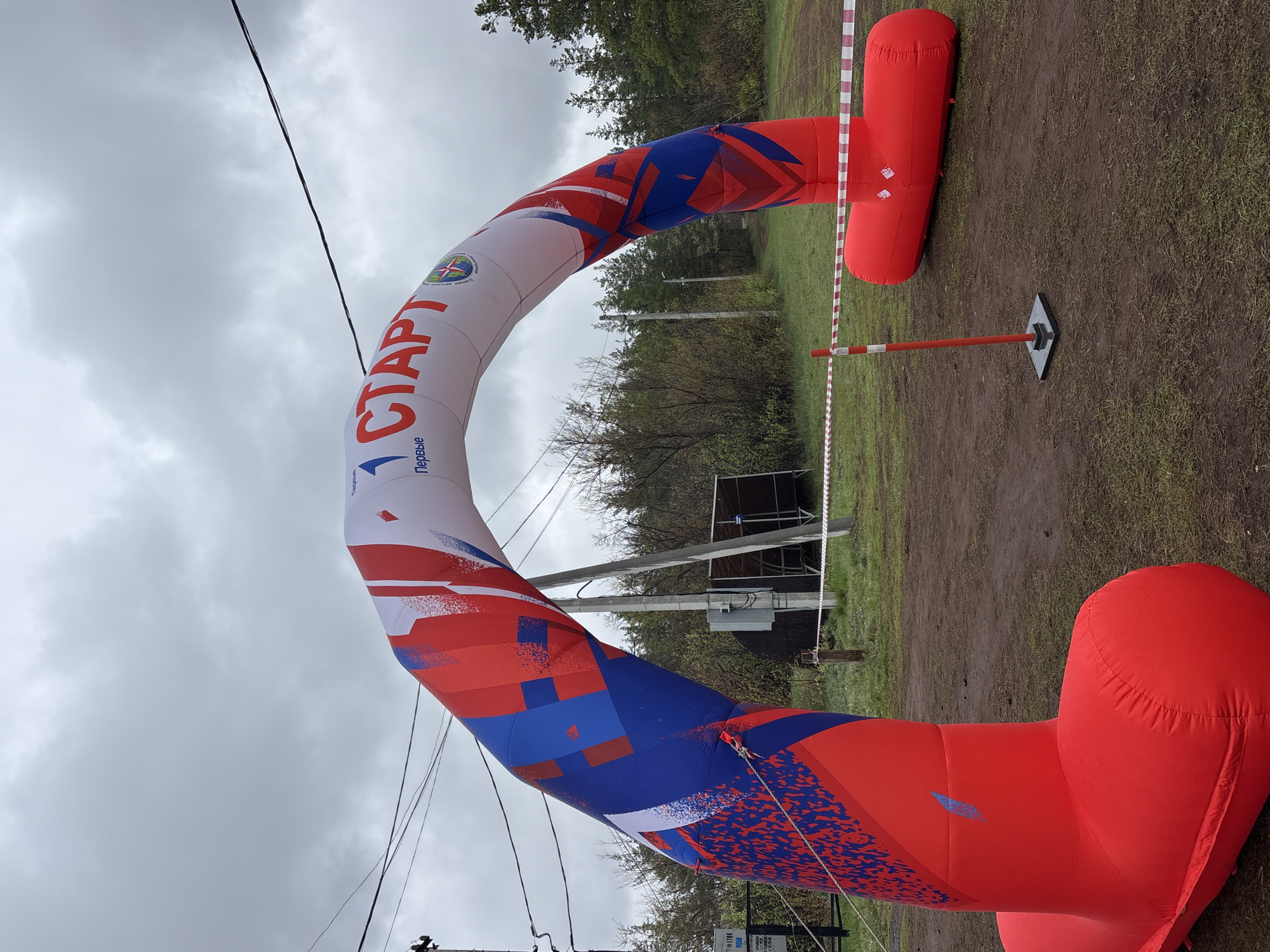 The image shows an inflatable archway marking the "START" of an event. The arch is primarily white with red and blue accents, and features the Russian word "СТАРТ" (Start) printed vertically. The archway is supported by large, red inflatable bases on either side. A barricade tape stretches across the background, and a support pole with a black base is visible to the right. The background includes trees and a structure that appears to be a small stage or shelter. Based on the metadata, the location is Orenburg, Russia. The sky is overcast, suggesting a cloudy day. The ground is a mix of dirt and grass. There are power lines visible in the upper left of the frame.