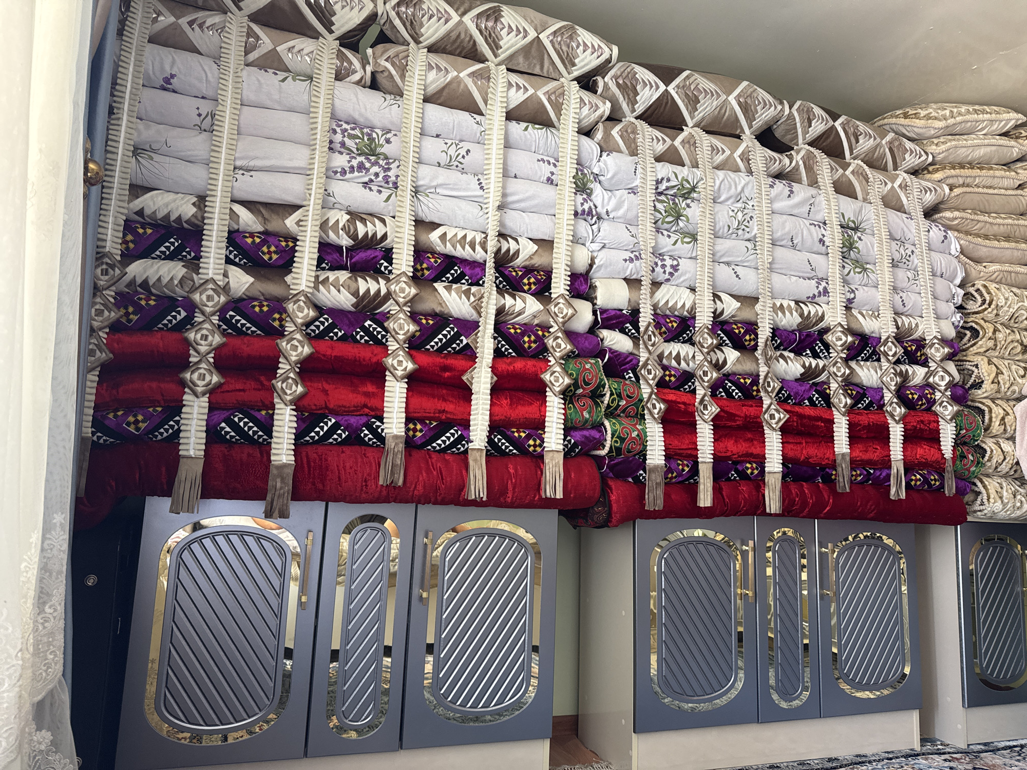 A vertically oriented, medium shot captures a stacked collection of pillows and bedding in Sovet, Kyrgyzstan, filling a significant portion of the frame. The items are organized in neat rows, with some featuring intricate patterns of purple, red, and geometric designs, while others are plain white with floral motifs. Decorative tassels and woven elements hang between the rows, adding a handcrafted touch. Below the bedding, two dark gray cabinets with ornate gold trim and ribbed detailing are visible. The left cabinet features a door and a narrow panel, while the right cabinet has two narrow panels. A portion of a white lace curtain hangs from the left edge of the frame, and a dark doorway is partially visible behind it. The background is a plain, light-colored wall. The lighting is even, suggesting an indoor setting, possibly a room dedicated to storage or display of traditional textiles.