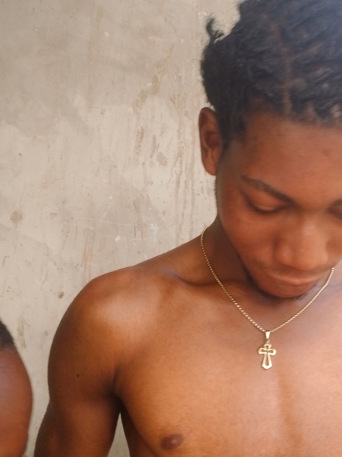 The image depicts a close-up of a bare-chested individual with medium-dark skin, observed in Enugwu-Ukwu, Nigeria. The subject's head is tilted downwards and to the right, with eyes directed away from the viewer. Dark hair is styled in braids or twists, pulled back from the face. A thin, gold-colored chain with a stylized cross pendant hangs around the subject's neck, resting on the upper chest. The left nipple is visible. A portion of a second individual's dark-skinned shoulder or upper arm is partially visible on the far left side of the frame, suggesting proximity. The background consists of a textured, light-colored wall exhibiting discolorations and streaks, consistent with weathering.