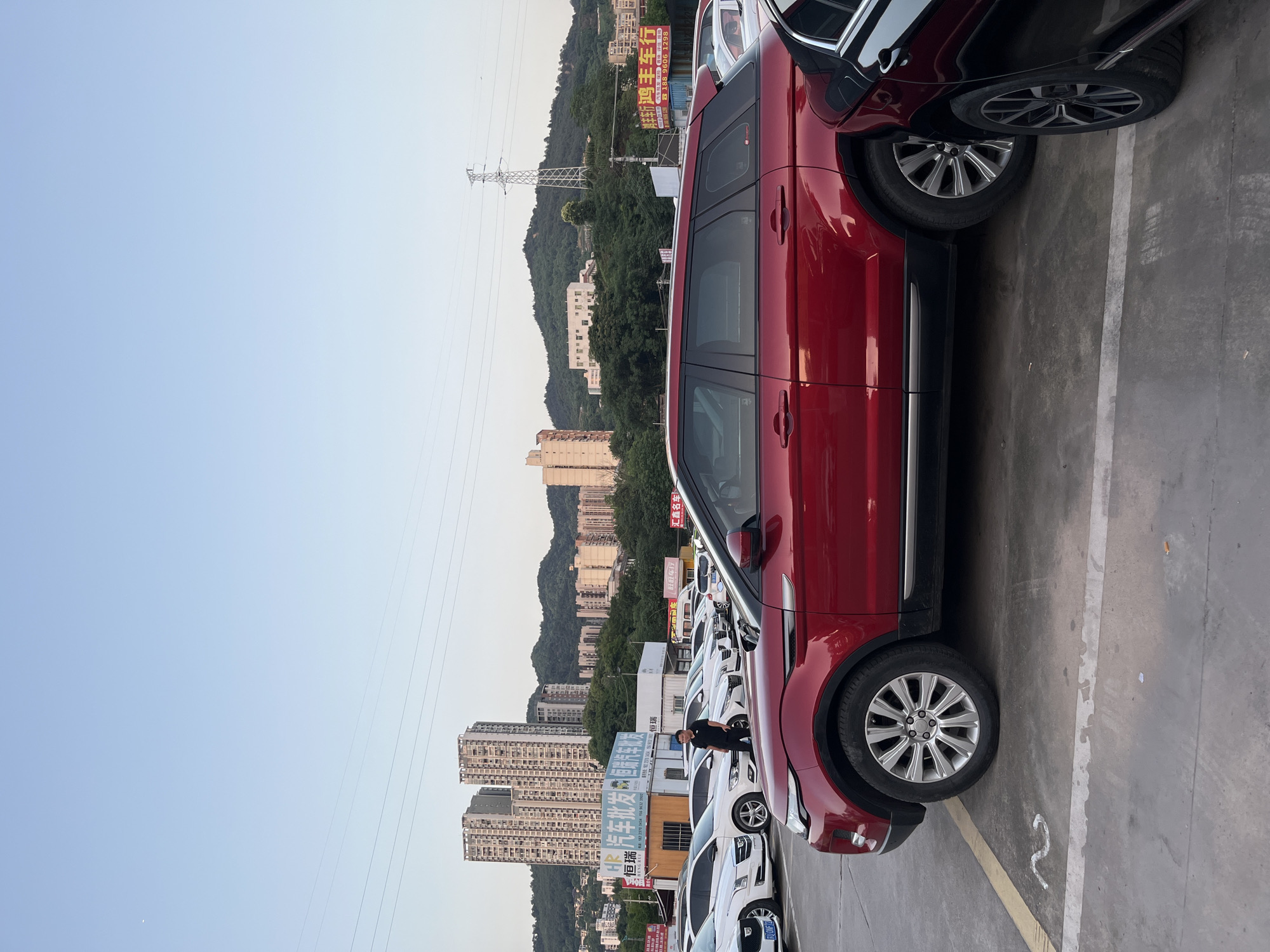 The image shows a parking lot in Banan, China, filled with cars. A red SUV occupies the foreground. Behind it, several white cars are parked in rows. In the background, high-rise buildings and tree-covered hills rise toward the sky. Power lines run across the scene. One man is standing near the cars. Signs with Chinese characters are visible above the parked cars.