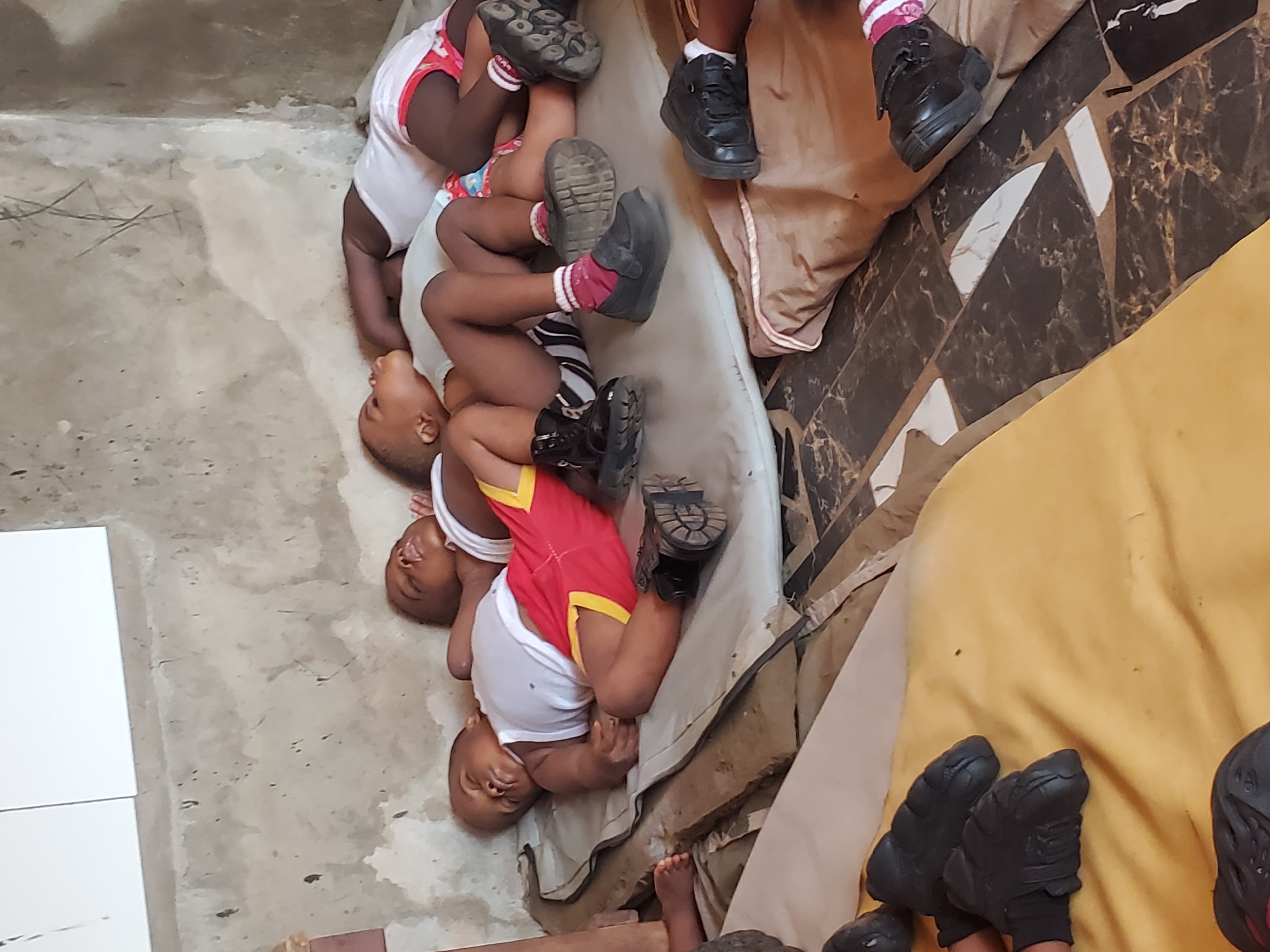 In Onitsha, Nigeria, three young children are lying on a thin mattress on the floor. The children are wearing white tank tops and shorts. One child has red shorts with yellow trim. The other two children have red and white shorts. The floor appears to be concrete. The mattress is placed near another mattress and other objects covered in fabric. The children are wearing shoes. One of the children has a bare foot.
