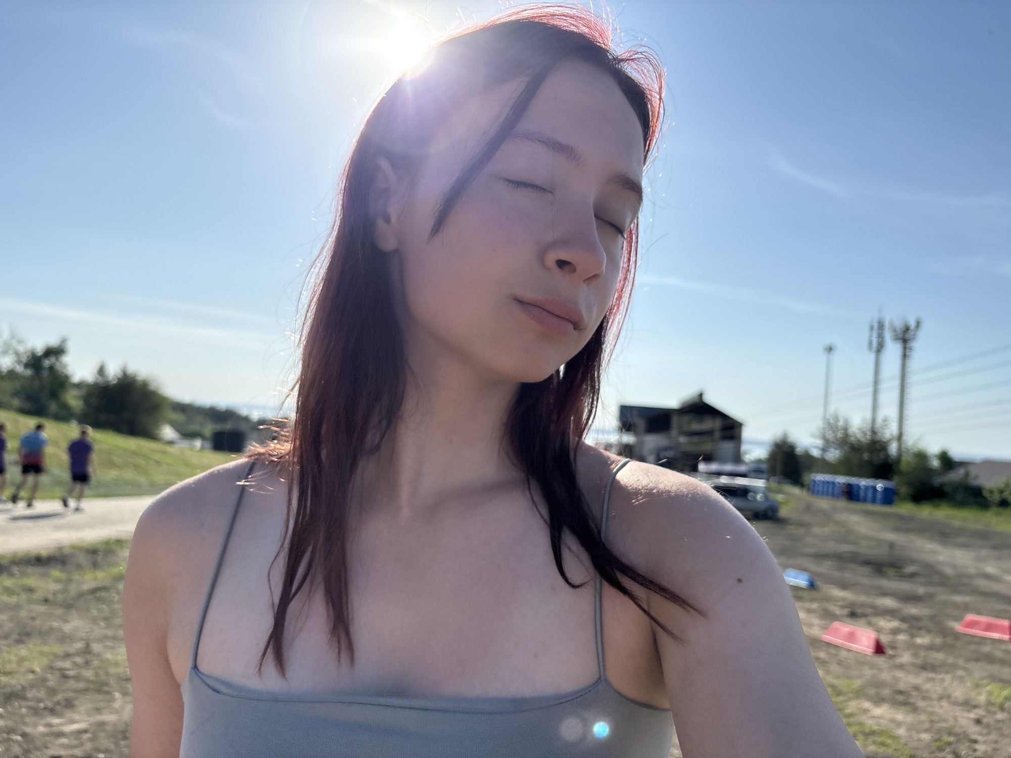 A young woman with reddish-brown hair and a light complexion stands outside with her eyes closed. She is wearing a grey tank top with thin straps. The sun shines brightly behind her head, causing a lens flare. In the background, a grassy hill is visible to the left, where three figures are walking. To the right, there is a building, some vehicles, and a row of portable blue toilets. The ground appears to be dirt or gravel. This image may be taken in Khvalynsk, Russia.