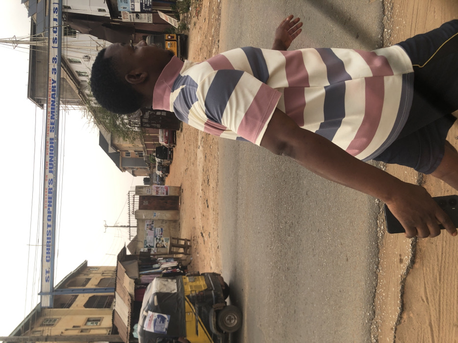 The image captures an outdoor urban or semi-urban scene in Akpaka Forest Reser, Nigeria, under an overcast or hazy sky. In the foreground, a dark-skinned male subject, with short dark hair, is walking from right to left, mid-stride. He is dressed in a short-sleeved collared shirt featuring horizontal stripes of white, light pink, and light blue, paired with dark shorts. A black mobile phone is held in his left hand, partially obscured by his leg.
In the midground, an overhead blue archway sign is prominent, displaying the text "DIOCESE ON THE NIGER (ANGLICAN COMMUNION) ST. CHRISTOPHER'S JUNIOR SEMINARY 3-3. (S.C.J.S.)". To the left of the paved road, a yellow and black motorized tricycle is partially visible, parked near a makeshift market stall with a corrugated metal roof and various items of clothing displayed. A wooden utility pole stands near the center of the image.
Beyond an open brown metal gate on the right, a dirt road extends into the background, where multiple vehicles are parked or visible. Multi-story buildings line both sides of the street, including a light-colored structure on the far left and a building with visible scaffolding on the right, behind the archway sign. The immediate ground consists of an asphalt road with a dirt shoulder to the right.