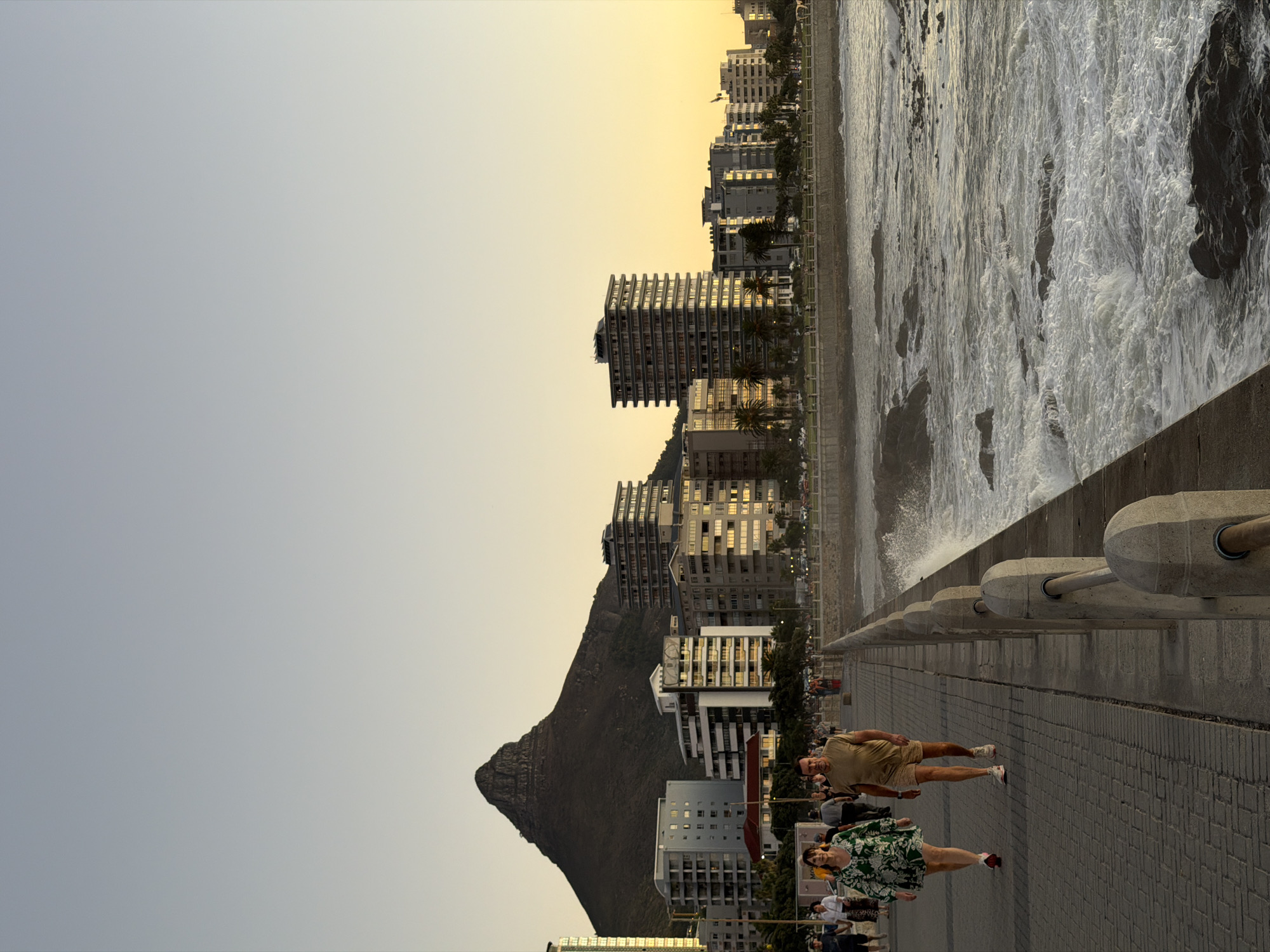 The image shows a landscape of Cape Town, South Africa, featuring Lion's Head mountain, buildings along the coastline, and the ocean. A wide sidewalk runs along the coast, bordered by a railing. People are walking along the sidewalk. The sky is light with a golden hue, indicating either sunrise or sunset. The ocean is turbulent, with white foamy waves crashing against the shore. Buildings of varying heights and architectural styles form the city's skyline. Lion's Head mountain looms in the background.