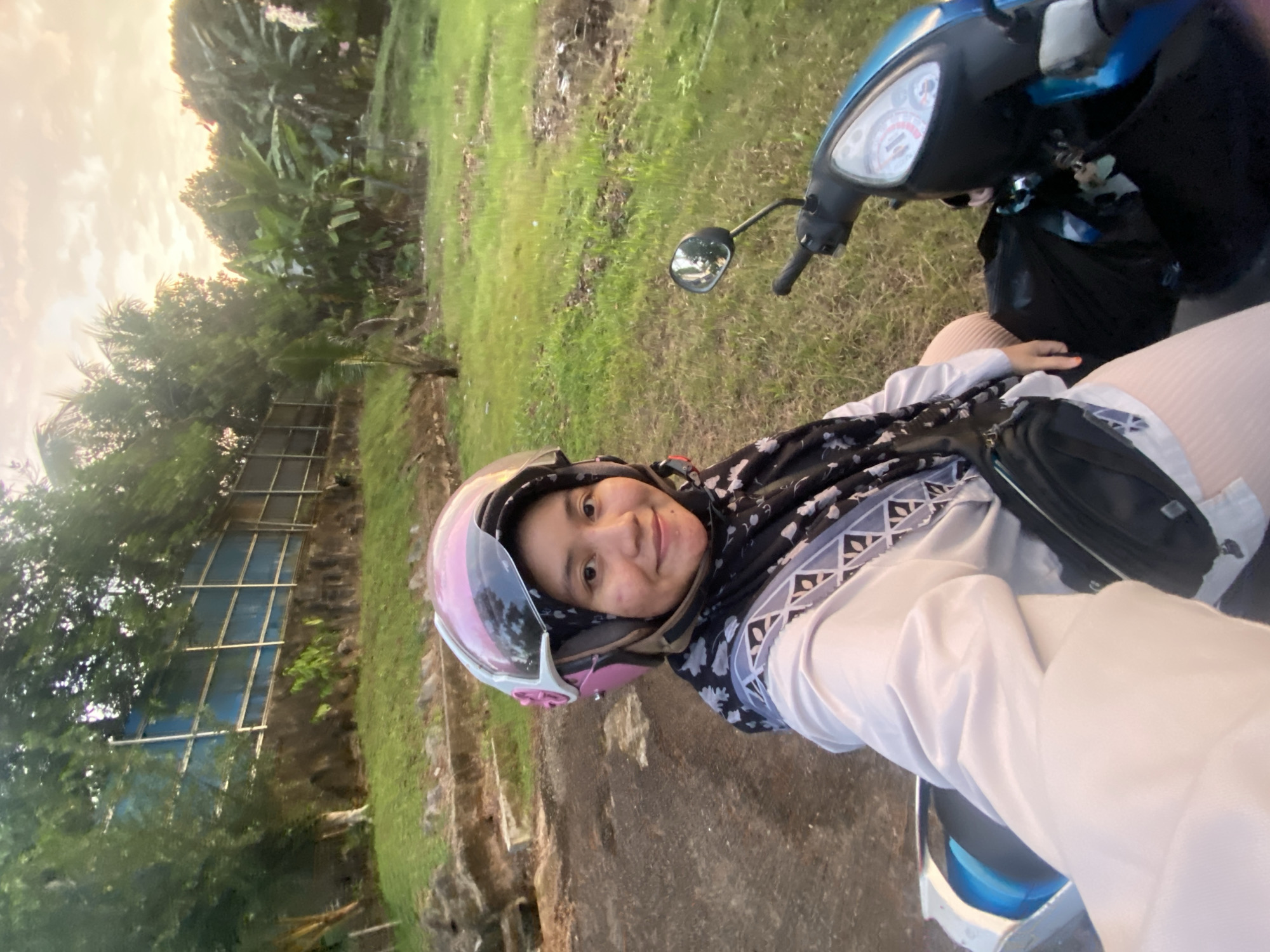 A woman is sitting on a blue and black motorbike. She is wearing a light pink helmet with a reflective visor, a black and white patterned headscarf, and a light-colored long-sleeved shirt. The woman is smiling and looking at the camera. She is positioned in the center of the frame, and the motorbike's handlebars and front are visible to her right. A black bag is attached to the motorbike's front.
The setting appears to be outdoors, with lush green grass and trees in the background. The sky is visible in the upper left corner, suggesting it is either early morning or late afternoon, with a warm, hazy light. There is a fence or structure made of metal bars and blue material visible behind some trees on the left. The ground where the motorbike is parked is a mix of dirt and asphalt.
The image is taken in portrait orientation and appears to be a selfie. The overall mood of the image is cheerful and casual. No visible text is present in the image. The context is Banjarbaru, Indonesia.