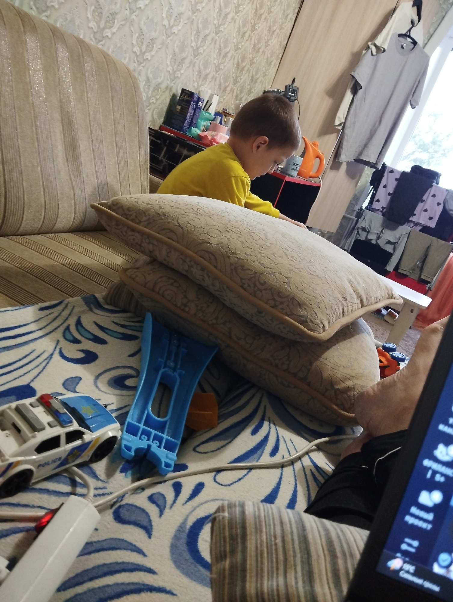 A young boy in a yellow shirt sits on a couch in a room. He is looking down at something out of frame. In front of the couch, on a patterned blanket, are a toy police car, a blue plastic toy, and some other toys. Two pillows are stacked on the couch next to the boy. In the background, there are clothes hanging on a rack and a table with items on it. The room appears to be a living room or play area. The lighting suggests it is daytime. There are no clear indicators of weather or specific emotions from the boy. Visible text includes "POLICE" on the toy car and some text on a device in the foreground, which is too blurry to read. The context provided is Kingisepp, Russia.