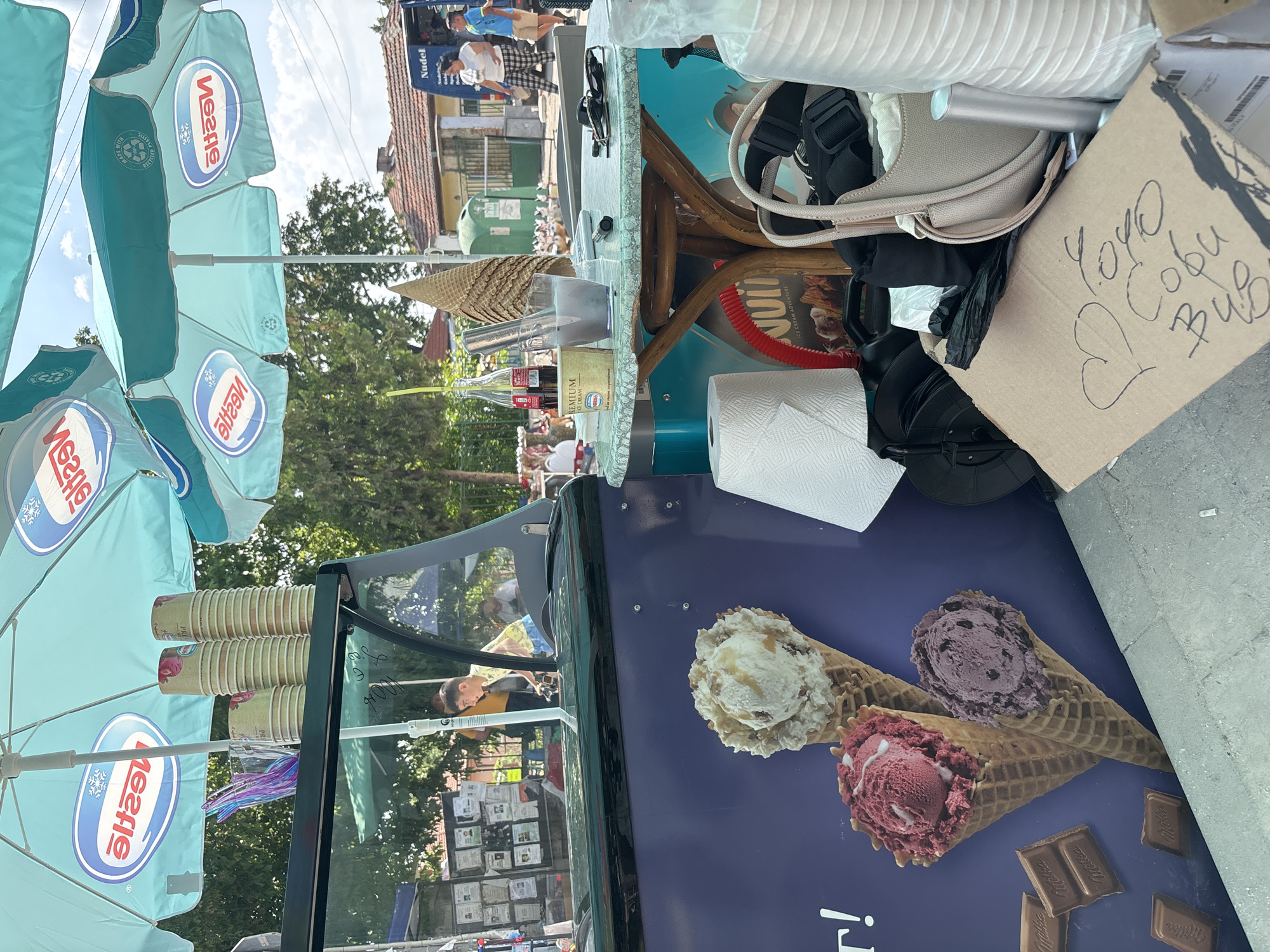 This image depicts an outdoor ice cream stand operating under large, light blue and green umbrellas branded with the reversed Nestle logo and a recycling emblem, indicating "MADE WITH RECYCLED PLASTIC." The scene is set in what is identified as Kostievo, Bulgaria, during daylight hours, characterized by bright sunlight and visible trees.
In the foreground, a dark green and blue mobile ice cream cart is positioned. Its visible side features a graphic displaying three ice cream cones: one with a light-colored scoop (possibly vanilla/caramel), one with a red scoop (berry-flavored), and one with a purple scoop (grape/chocolate). Several squares of Milka chocolate bars are also depicted on the graphic. On a shelf above the cart, stacks of paper cups with a fruit design and a clear glass containing colorful plastic spoons are visible.
Adjacent to the cart, a small, round table with a mottled light green surface and wooden legs holds various items: a stack of ice cream cones, two red Coca-Cola bottles, a glass containing a metal ice cream scoop or stirrer, a white "PREMIUM ICE CREAM NESTLE" tub with a purple lid, sunglasses, and a dark pen or lighter.
On the ground next to the table, several objects are present: a roll of white paper towels, a red corrugated hose, a beige handbag with black straps, other dark fabric items, stacks of white paper cups, and a cardboard box. Handwritten Cyrillic script, possibly "Yoyo Софи Бул" or similar, is present on the cardboard box. A black plastic object is partially visible beneath the table.
In the mid-ground, a young boy in a yellow and blue patterned shirt is visible behind the counter of the ice cream cart, looking right. A second person in a yellow shirt is partially obscured further behind the counter. To the left of the boy, a wall covered with numerous posted notices or flyers is visible.
The background shows a street scene with older, single-story houses featuring tiled roofs. A blue commercial van, partially labeled "Nudel" on its side, is parked on the street. Near the van, a green public waste bin is visible. Two individuals are walking away from the viewer on the street: a woman in a white t-shirt and checkered pants, and a man in a blue t-shirt and beige shorts. The general environment includes ample green foliage from trees and a partly cloudy sky. The ground consists of paved surfaces.