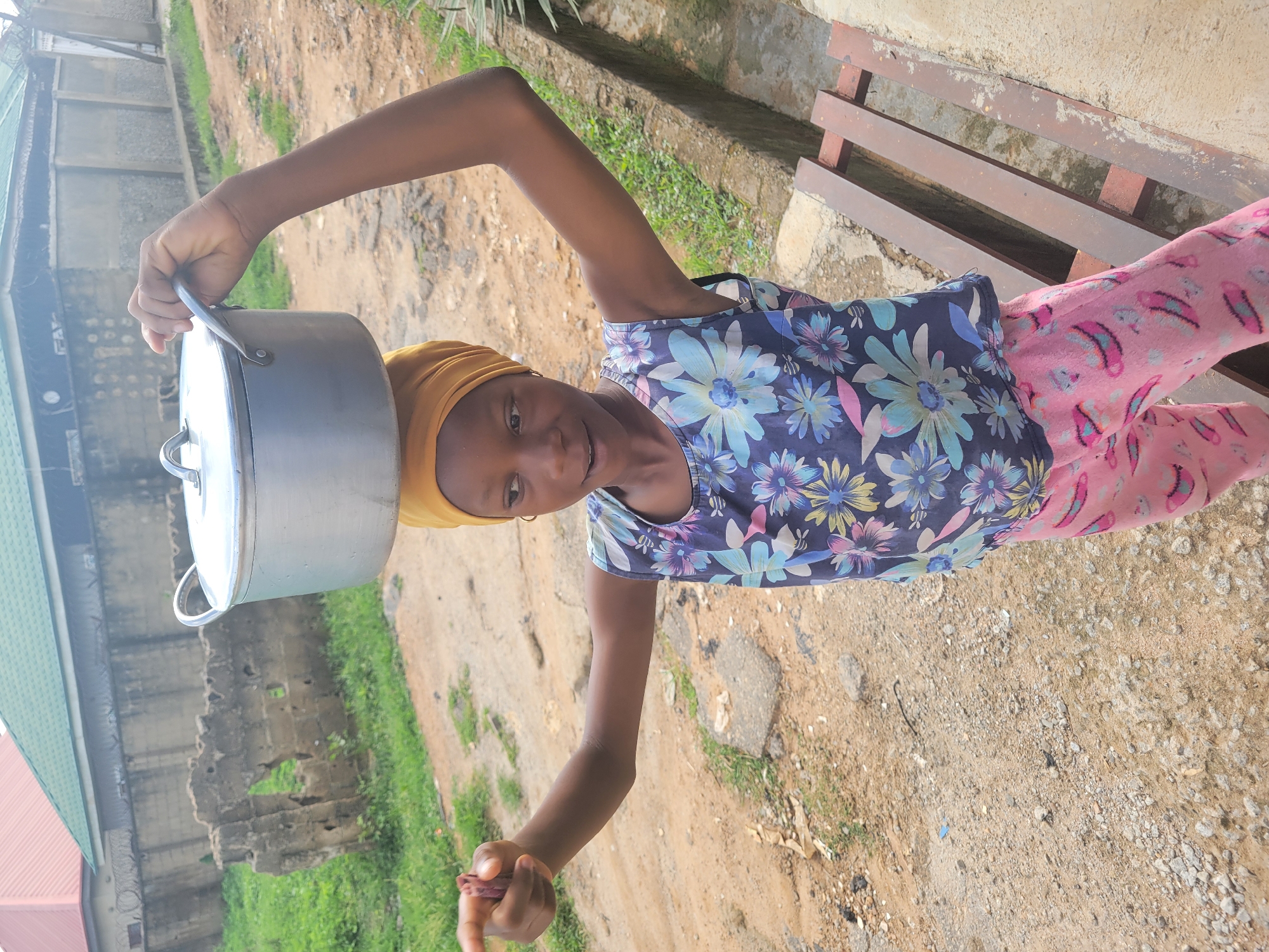 A young woman, likely a teenager, stands on a dirt and gravel surface in what appears to be an outdoor environment. She is wearing a yellow head covering, a floral print sleeveless top, and pink patterned pants. She balances a large, silver cooking pot on her head with one hand. Her other hand holds a small, reddish-brown object. Behind her, there is a dilapidated wall with a building and green vegetation visible. To her right, there is a wooden bench against a light-colored wall. The location is Madalla, Nigeria.