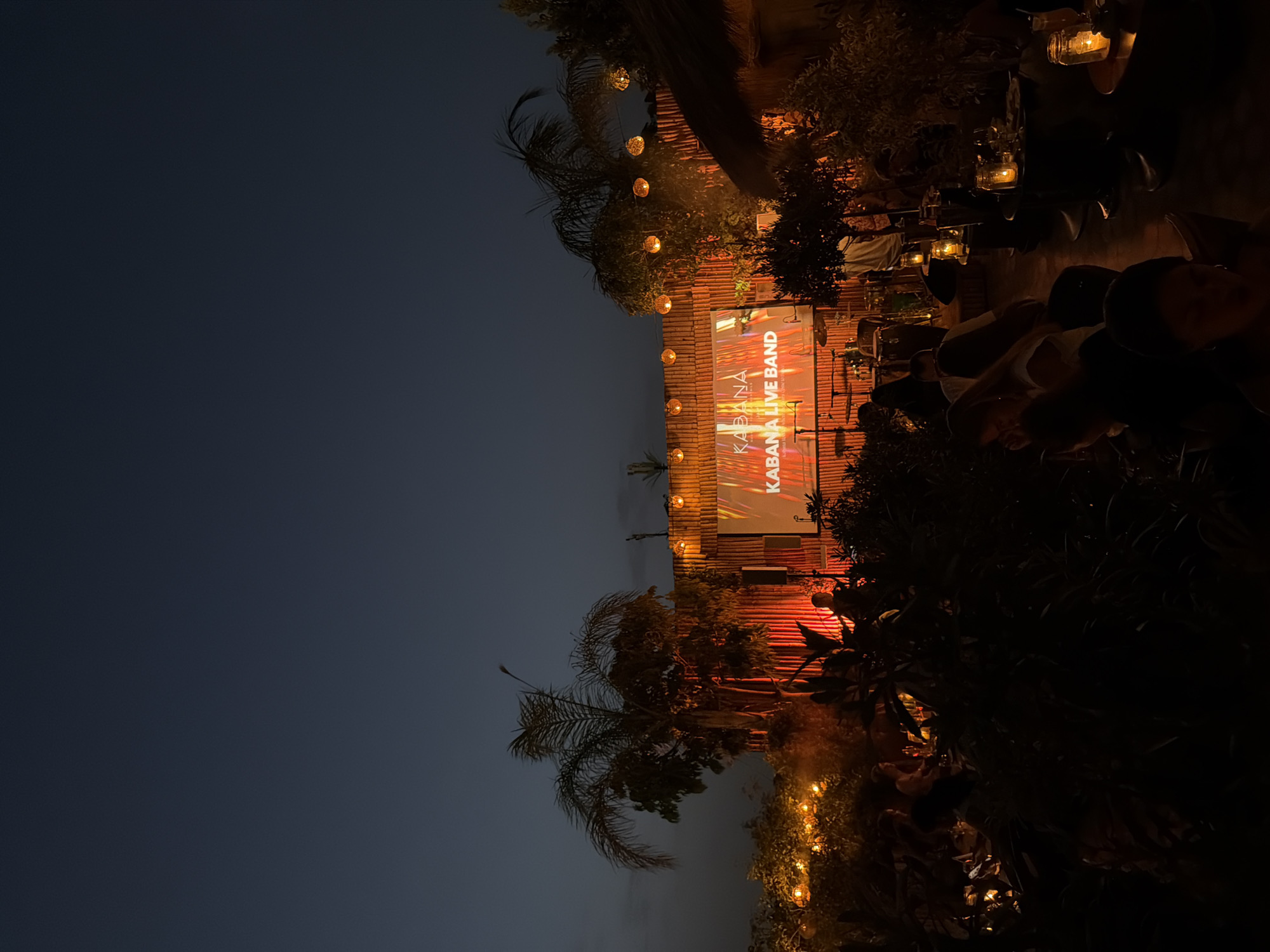 The image depicts an outdoor evening scene at Kabana Marrakech, a venue in Marrakesh, Morocco. The sky is a deep twilight blue. In the background, a bamboo-clad stage is illuminated with warm orange lighting, featuring a large screen displaying "KABANA LIVE BAND." Several palm trees are visible against the sky. Strings of ambient yellow globe lights hang across the scene, casting a warm glow. In the foreground, clusters of people are seated at tables, some illuminated by lanterns, creating a relaxed atmosphere. Lush greenery and tropical plants surround the seating areas. The overall impression is of a lively entertainment venue during an evening gathering.
