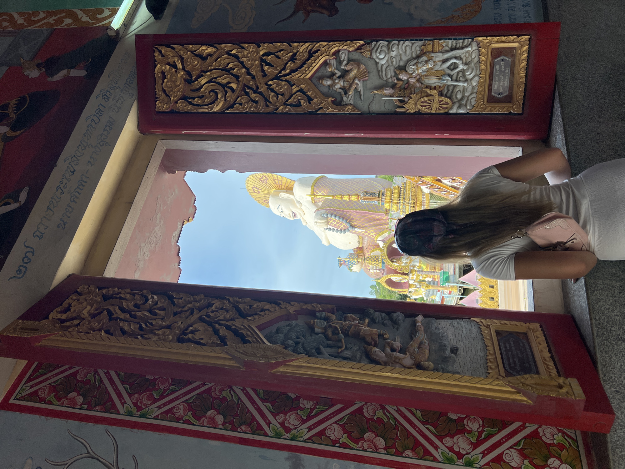 A woman with long blonde hair and a pink purse is standing at a window, looking out at a large Buddha statue in Baan Plai Laem, Thailand. The Buddha is white with a golden halo and is holding prayer beads. The window is framed by ornate red and gold carvings and paintings of flowers. The sky is blue.