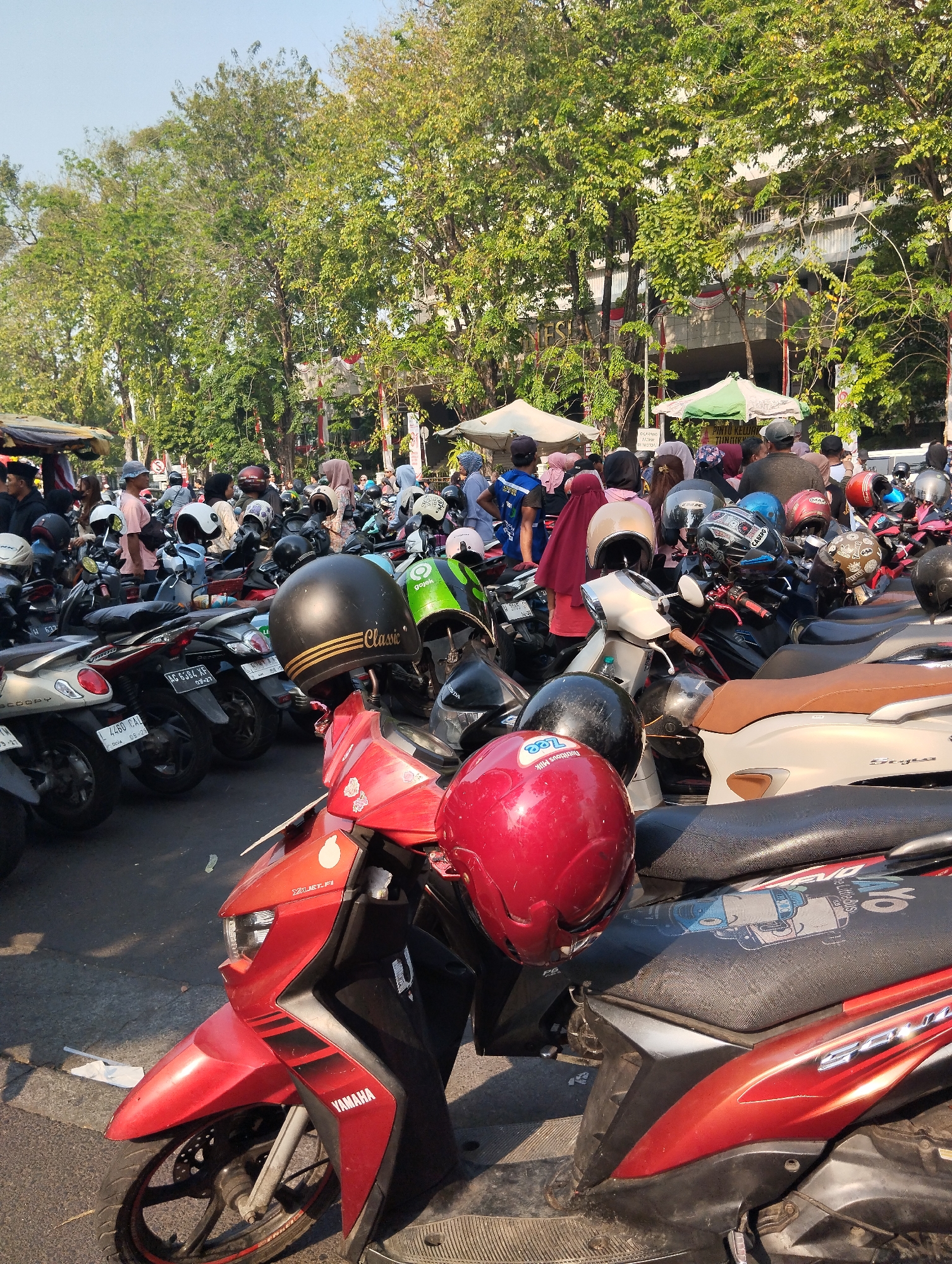 The image depicts a busy outdoor parking lot filled with numerous motorcycles in Surabaya, Indonesia. The lot is adjacent to a building with trees lining the foreground. In the foreground, a red motorcycle with the "YAMAHA" logo is prominently displayed, with a red helmet resting on its seat. Other motorcycles, of various colors and models, are parked closely together, with several helmets scattered amongst them. Many people are visible in the background, some walking and others standing near the motorcycles. Tents are set up in the background, suggesting a market or event. The overall scene is one of a vibrant and active urban environment.