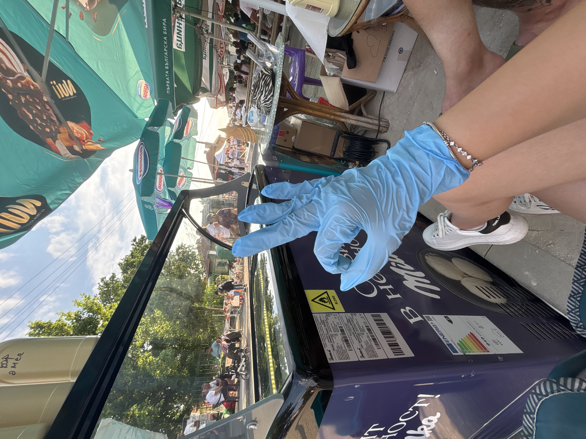 The image depicts an outdoor daytime scene in Kostievo, Bulgaria, likely an open-air cafe or market area. In the immediate foreground, a person's left hand, covered by a light blue disposable glove, forms an "OK" gesture. A silver-colored bracelet with small white stones is visible on the wrist. To the left, a portion of a white sneaker with a black heel tab is present, while to the right, a bare foot with a tattoo on the lower leg is visible.
A dark blue commercial display freezer occupies the left side of the foreground. Its reflective glass top shows an inverted reflection of the background. The freezer features an EU energy label displaying "Class D" and "949 kWh/annum." A warning symbol for flammable R290 refrigerant is present, along with Cyrillic text identifying it as a "JUKA Model: Display freezer R440SL." "Milka" branding is visible on the side, accompanied by an image of round items, possibly pancakes or cookies. Partial text "IT HOCT!" and "lka" is also visible. Above the freezer, partially inverted Cyrillic text "вар а мен" is discernible, possibly from a menu board.
The midground and background reveal a bustling outdoor environment. Multiple people are seated at tables under large commercial umbrellas. These umbrellas display various branding, including "Nestlé," "Milka" (with an image of a chocolate ice cream bar), and "SLUNCHO," along with the Cyrillic text "ПЪРВАТА БЪЛГАРСКА БИРА" (The First Bulgarian Beer). Items on the tables include a stack of ice cream cones, plastic cups, and a zebra-patterned bag. Underneath the tables, boxes and other objects are stored, including one marked "FRAGILISSIMO." Several individuals are walking or standing in the background, and a child's tricycle is situated on the ground. Green trees, a wooden fence, and distant buildings with red-tiled roofs contribute to the backdrop. Overhead, power lines cross a blue sky with scattered white clouds, illuminated by sunlight that suggests late afternoon or early evening.