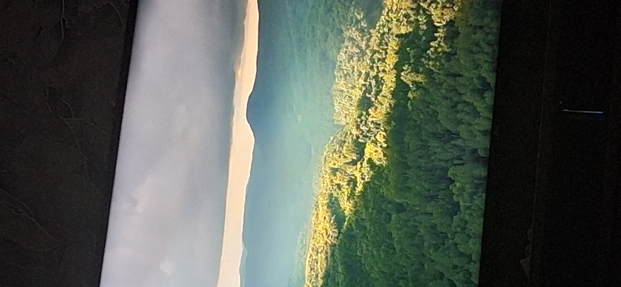 The image displays an aerial view of a densely forested area with rolling hills and distant mountains. Sunlight dramatically illuminates a section of the forest, creating a stark contrast with the shaded areas. A thin, bright band of light is visible between the clouds and the mountains, suggesting a sunrise or sunset. The lower portion of the image shows the dark frame of what appears to be a television screen. There are no people, animals, or man-made structures visible in the natural landscape. The provided crops focus on the top and bottom edges of the image, highlighting the dark frame and the transition to the sky, as well as close-ups of the forest canopy. There is no information within the image or its crops to suggest a location in Bila Tserkva, Ukraine.