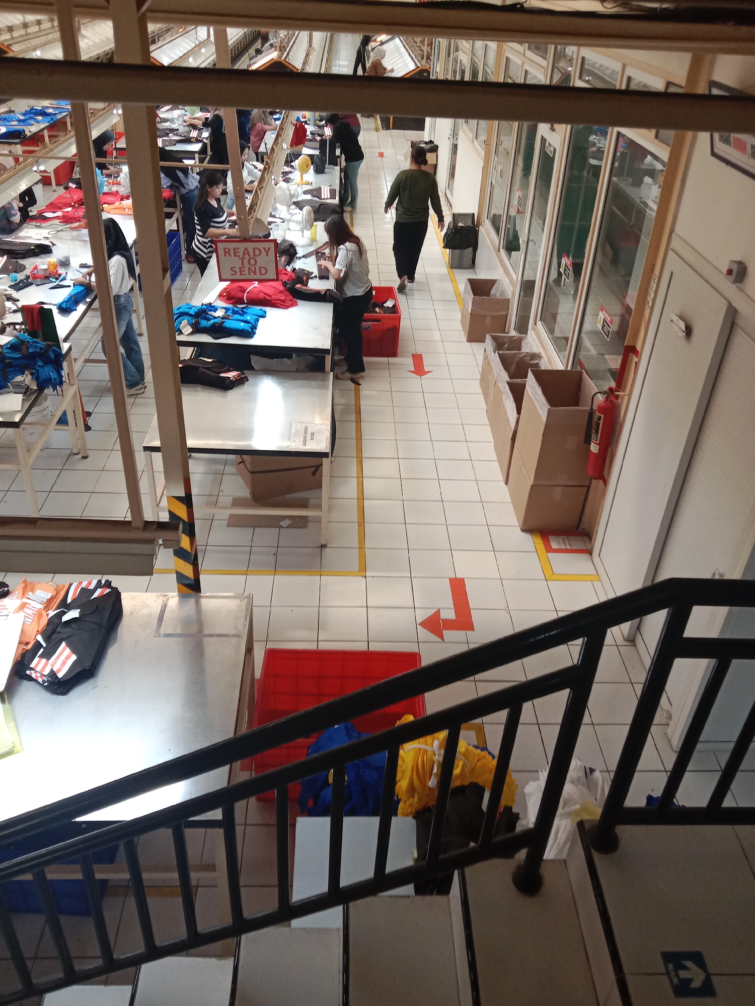 This image, taken in Semarang, Indonesia, depicts a busy garment factory floor viewed from a high angle, likely from a second-story walkway or stairs. The environment is a large, open space with tiled flooring marked with yellow and orange directional arrows. Rows of long, metal-topped work tables are visible, some laden with stacks of clothing, primarily t-shirts and workwear in various colors, including blue, red, black, and yellow.
Several individuals, predominantly women, are engaged in work at these tables. Some are wearing headscarves, indicating a Muslim-majority environment. One woman in a striped shirt is standing near a sign that reads "READY TO SEND." Other workers are seen interacting with the garments, folding them, or placing them in containers.
The space is organized with clear pathways, some marked with yellow lines. Various containers are present, including red plastic bins and cardboard boxes, some of which are stacked. Towards the right side of the image, a red fire extinguisher is mounted on a wall. Beyond the work area, there are glass-enclosed offices or sections, suggesting a structured production and administrative setup. The overall impression is one of an active manufacturing environment focused on textile production and distribution.