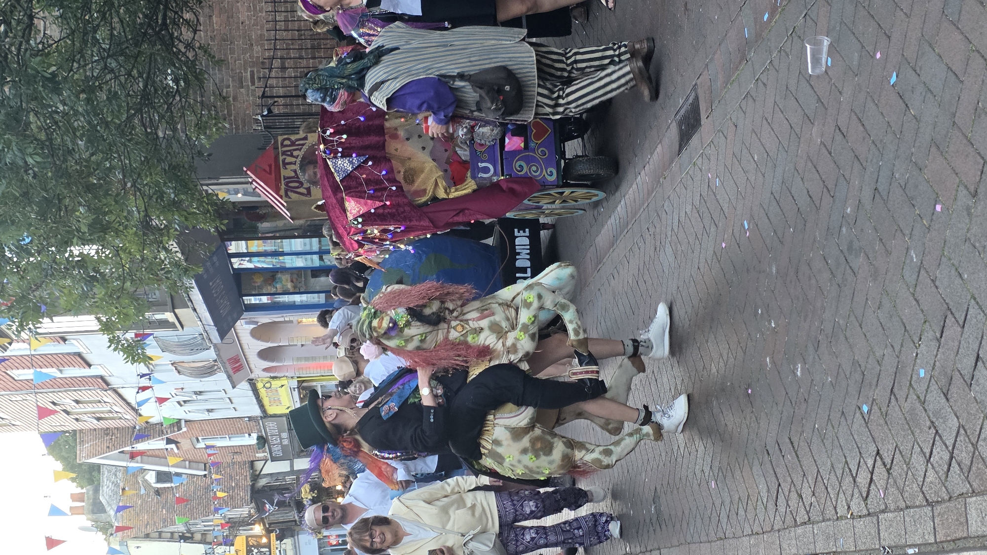 In St Leonards, United Kingdom, a person in a top hat and suit rides a horse costume on a cobblestone street, seemingly during a parade or festival. The horse costume is light-colored with green spots and has pink hair around its face. The rider is facing left, looking towards the camera. To the right of the rider, a decorated wagon with a velvet canopy and lights is being pushed by a person wearing striped trousers and a vest. The wagon has "ZOLTAR" written on a sign. Behind the wagon, a large globe is visible. The street is lined with buildings, some adorned with colorful flags. Confetti is scattered on the ground. Several other people are in the background, some observing the procession and others participating.