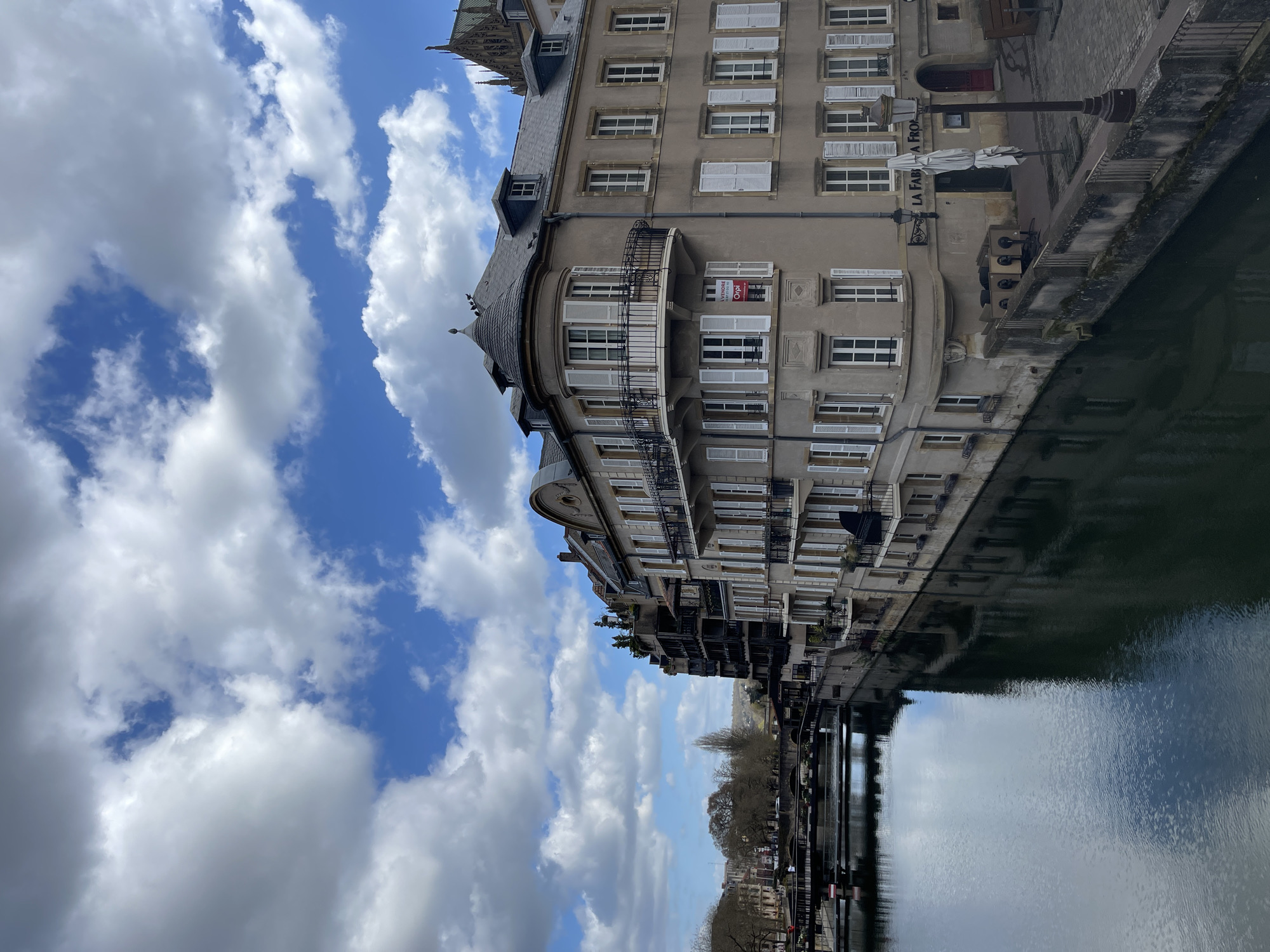 The image shows a building with a rounded corner and many windows with white shutters, located in Metz, France. It is likely near the Moselle river, as its reflection is visible in the water. The sky is visible with some white and grey clouds. The building appears to be multi-storied and has a balcony on the rounded corner. There is a sign that reads "LA FABRICA FROG". There is a red "A VENDRE" sign hanging on one of the windows.