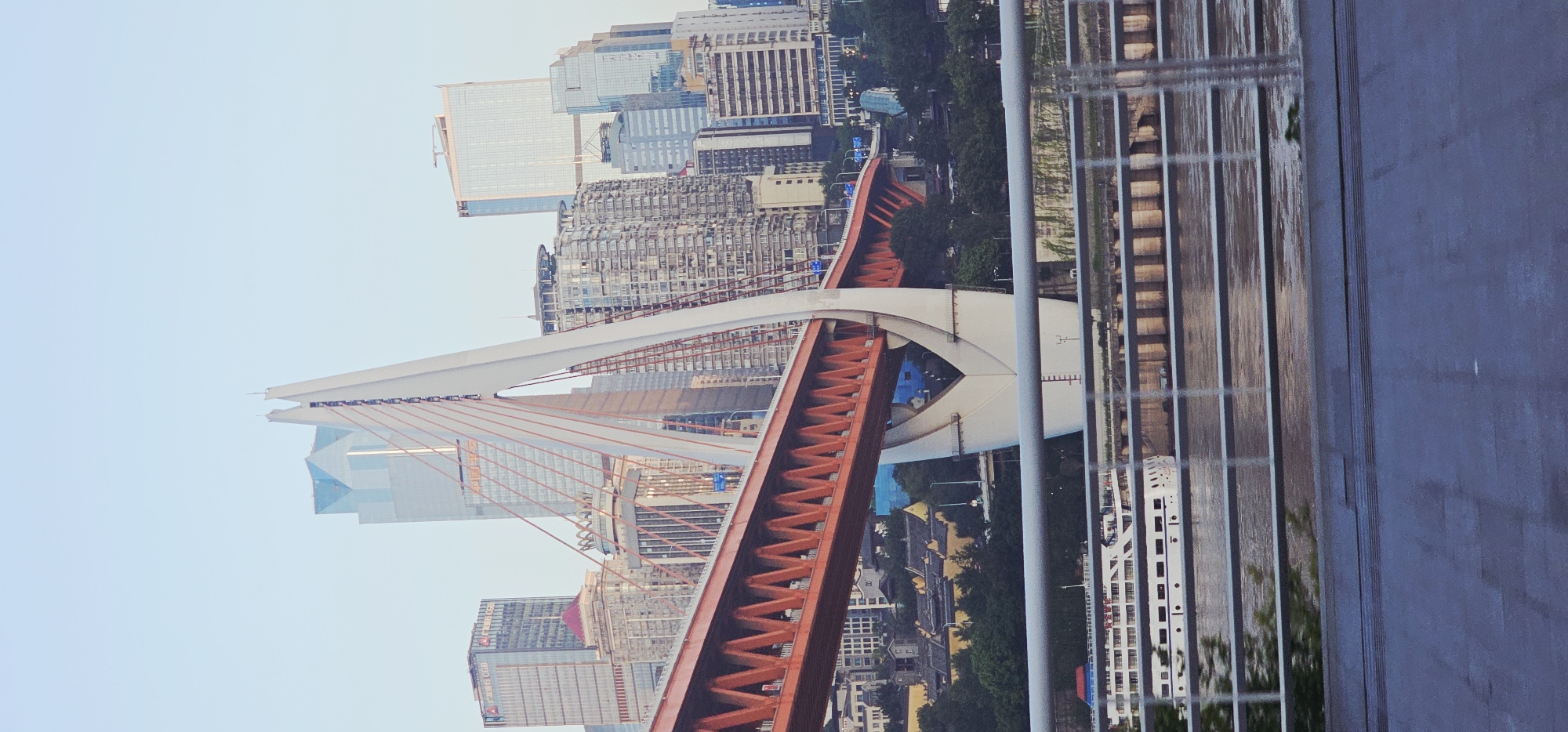 This is a vertical, eye-level, full shot of the Dongshui Gate Bridge in Nan'an District, China, taken from a moving vehicle at eye level, likely on the bridge or an adjacent road. The sky is a clear, pale blue.
The foreground is dominated by a grey, wet road surface with horizontal lines, suggesting it might be a street or highway. A silver railing with horizontal bars separates the road from the view beyond.
The middle ground features the river, with a white passenger ship docked or moving slowly. The ship has multiple decks and windows, and appears to be a large vessel. Beyond the river, the distinctive orange and white structure of the Dongshui Gate Bridge is prominent. The bridge has an orange truss section and a large white pylon with red suspension cables.
The background is a dense urban landscape of numerous modern skyscrapers with glass facades, rising behind the bridge and extending into the distance. Some buildings have a metallic sheen, reflecting the sky. The architecture is varied, with some buildings appearing taller and more contemporary than others. Green trees are visible between some of the buildings, adding a touch of nature to the cityscape. The overall impression is a modern city with significant infrastructure development.