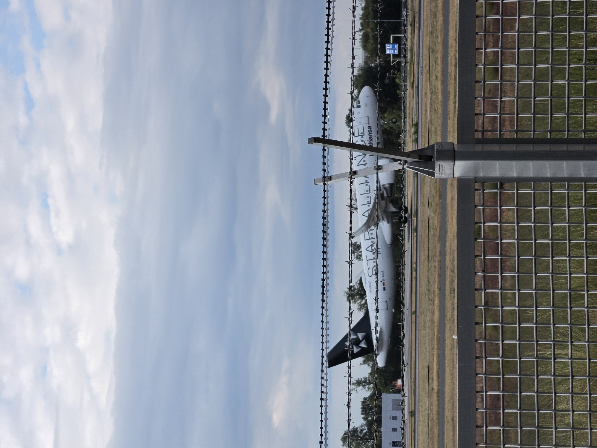 The image captures a Lufthansa airplane, identifiable by the Star Alliance logo and the text "Lufthansa" on its fuselage, on a runway. The plane has its landing gear deployed, suggesting it is either preparing to land or has just taken off. The scene is viewed through a barbed wire fence and a metal mesh fence, indicating it is taken from an airport perimeter. In the background, trees and airport buildings are visible under a cloudy sky. The location is the city of Kelsterbach, Germany, which is home to Frankfurt Airport, a major international hub.