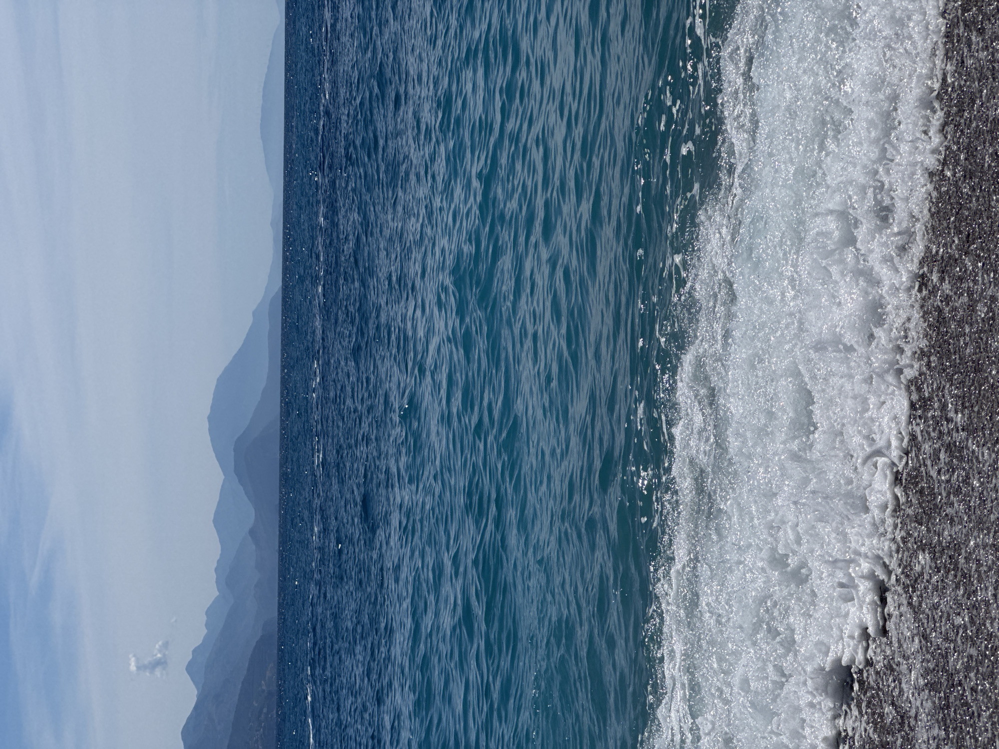 The image depicts a serene coastal scene with the Adriatic Sea in the foreground and a mountain range in the background. The water is a vibrant blue, with small waves gently lapping onto a dark, pebbly shore. In the distance, several layers of mountains fade into a hazy blue, suggesting a considerable distance. The sky is a pale blue with some wispy clouds. The overall impression is one of tranquility and natural beauty. This view is consistent with the landscape found in Prijevor, Montenegro, a region known for its stunning coastline and mountainous terrain. No people or specific structures are visible in the image.