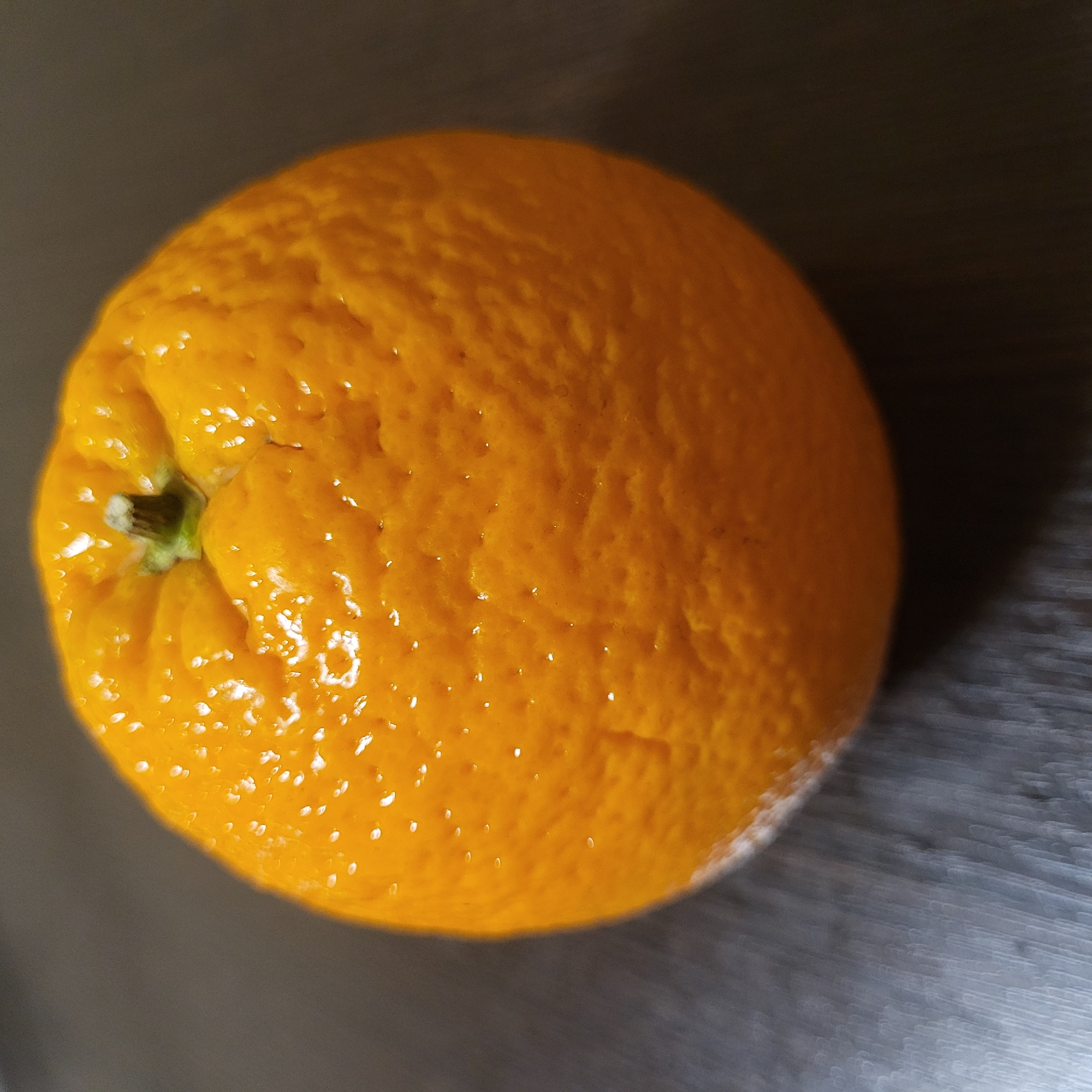An orange sits on a dark gray, possibly metallic surface. The orange is round, textured, and bright orange in color. It appears to be sitting still, casting a shadow on the surface. The scene is devoid of any other objects or people. Location and context are not discernible from this image. The location within Yerevan, Armenia is not indicated.